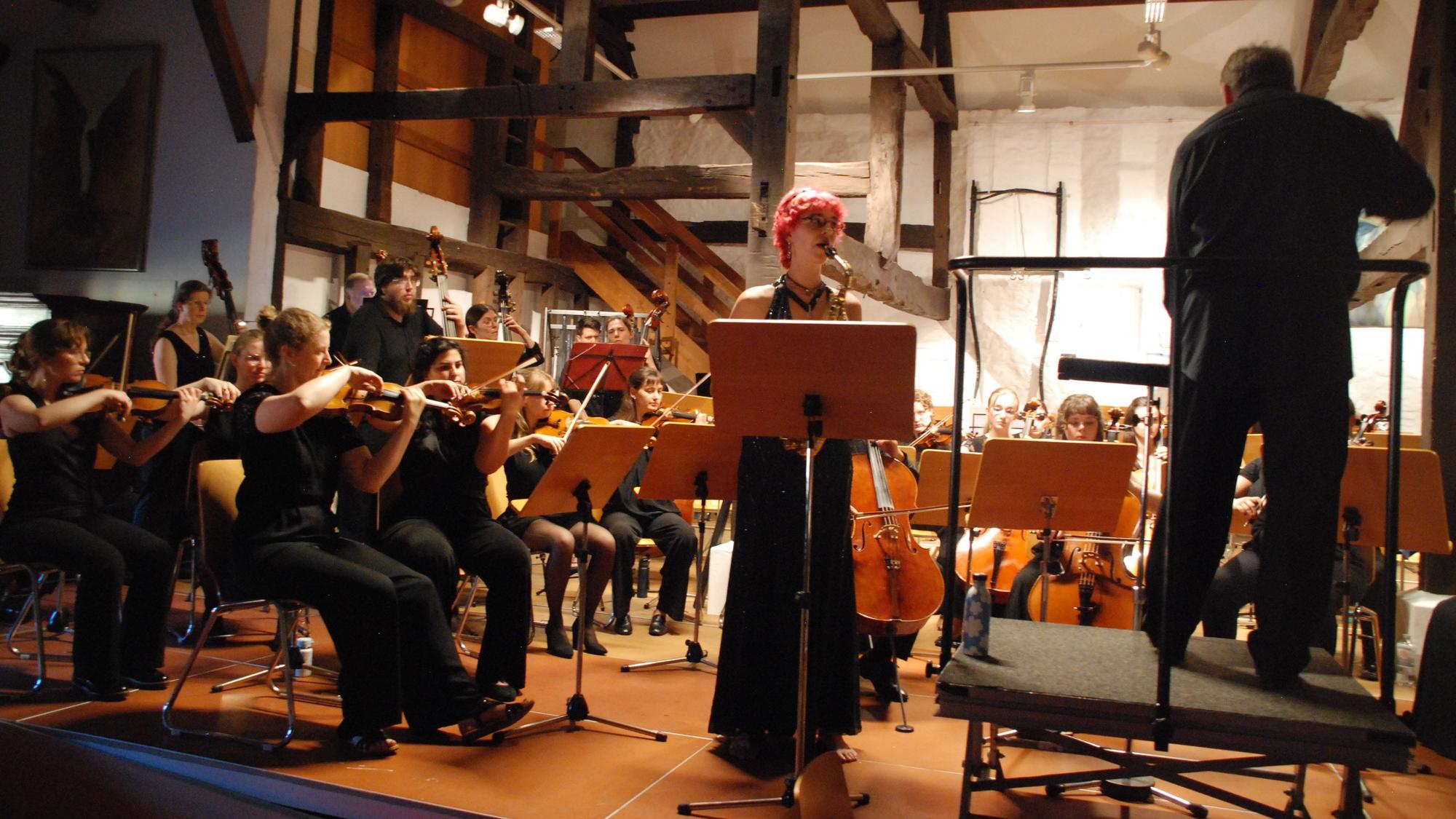 Lilian Eicks steht hinter einem Notenpult und spielt Saxofon. Links von ihr befindet sich das Orchester, rechts der Dirigent,.