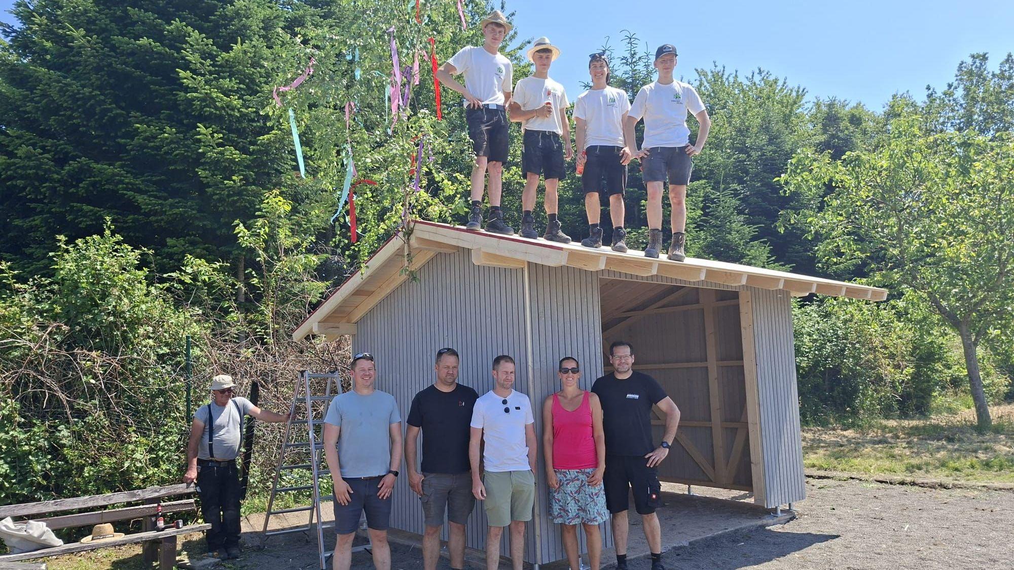 Zu sehen sind einige Menschen vor einer nicht ganz fertig gebauten Hütte im Grünen.