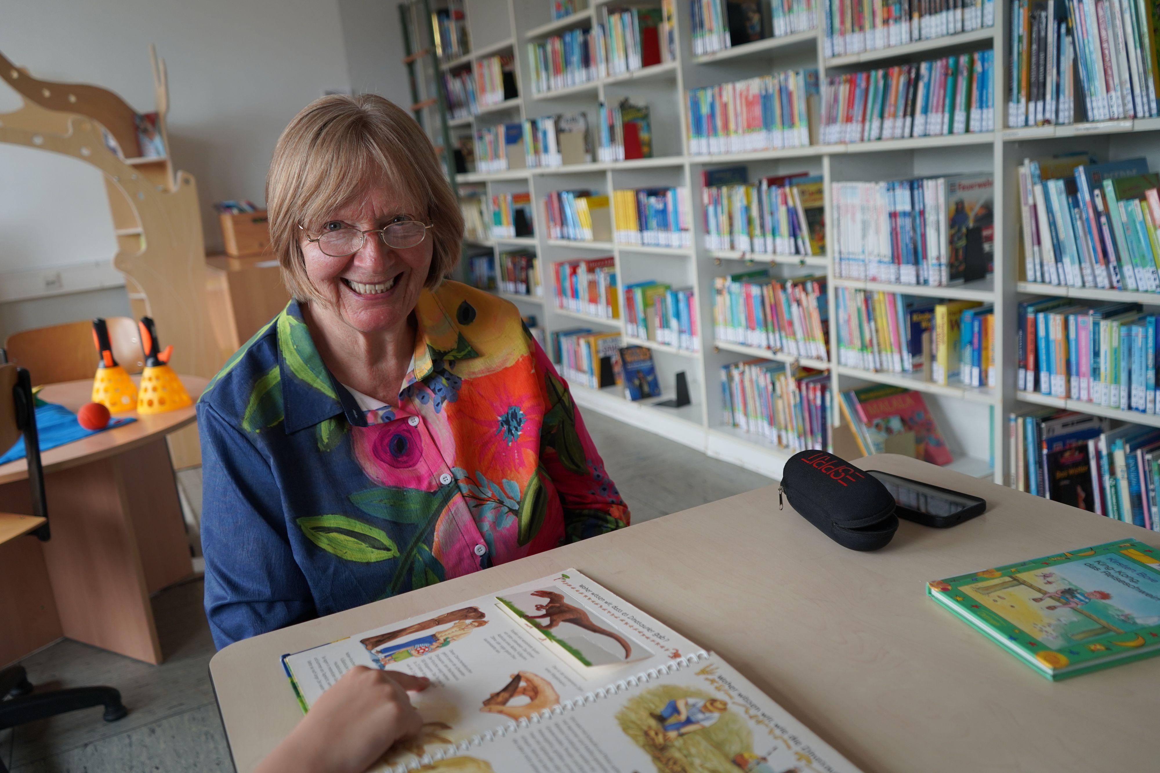 Das Bild zeigt die Seniorin im Lesewald der Rochusschule in Glessen. Vor ihr zeigt ein Kind gerade auf ein Buch.