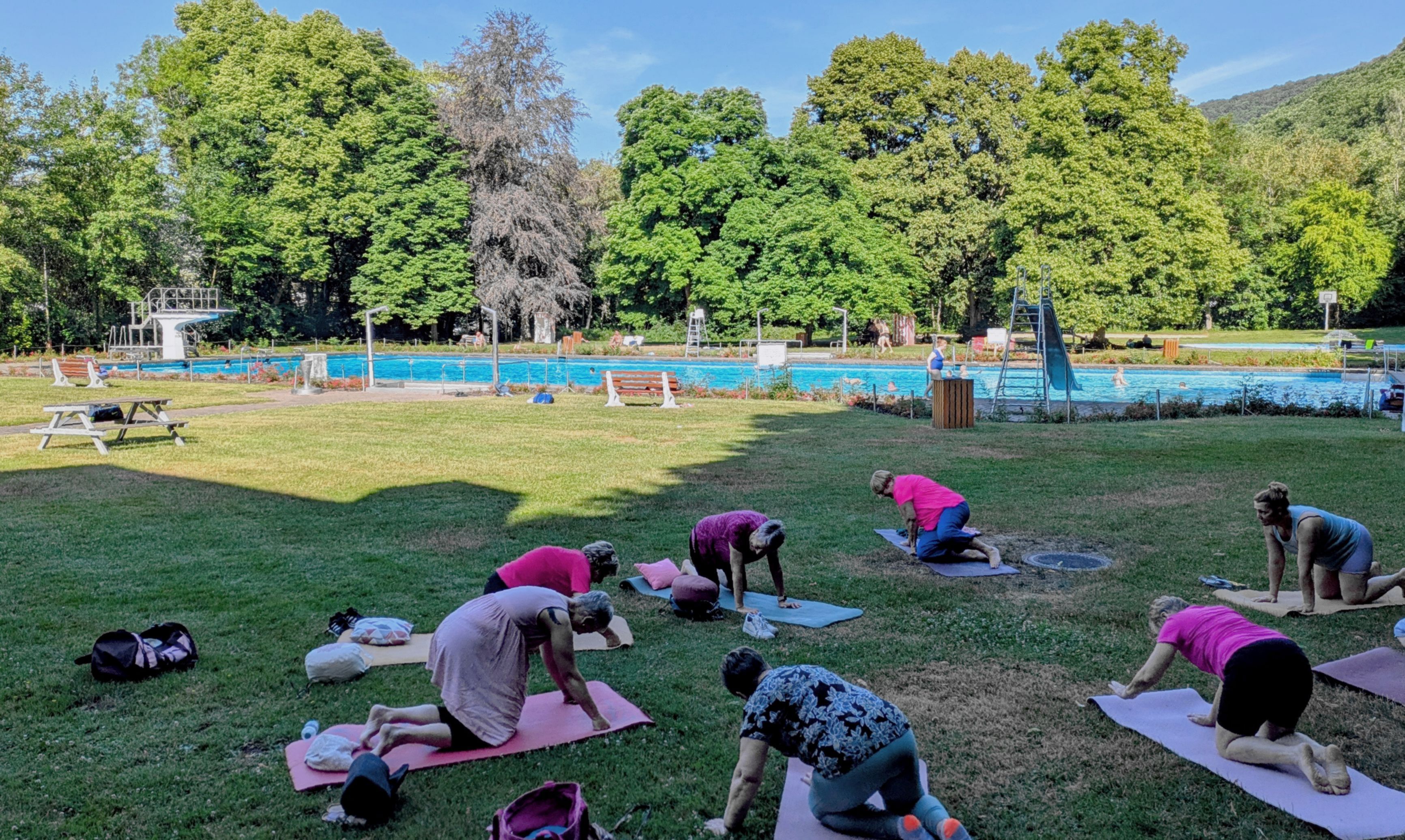 Auf der Liegewiese des Gemünder Rosenbads nehmen mehrere Frauen an einer Yogastunde teil. Im Hintergrund ist das Schwimmbecken zu sehen.