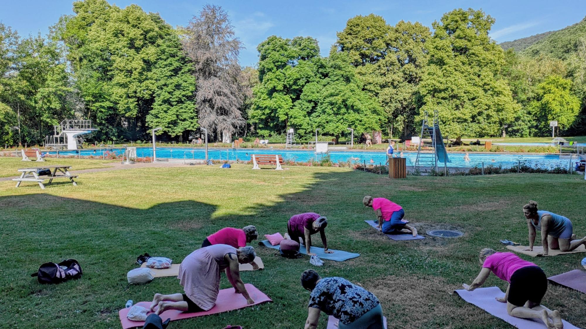 Auf der Liegewiese des Gemünder Rosenbads nehmen mehrere Frauen an einer Yogastunde teil. Im Hintergrund ist das Schwimmbecken zu sehen.