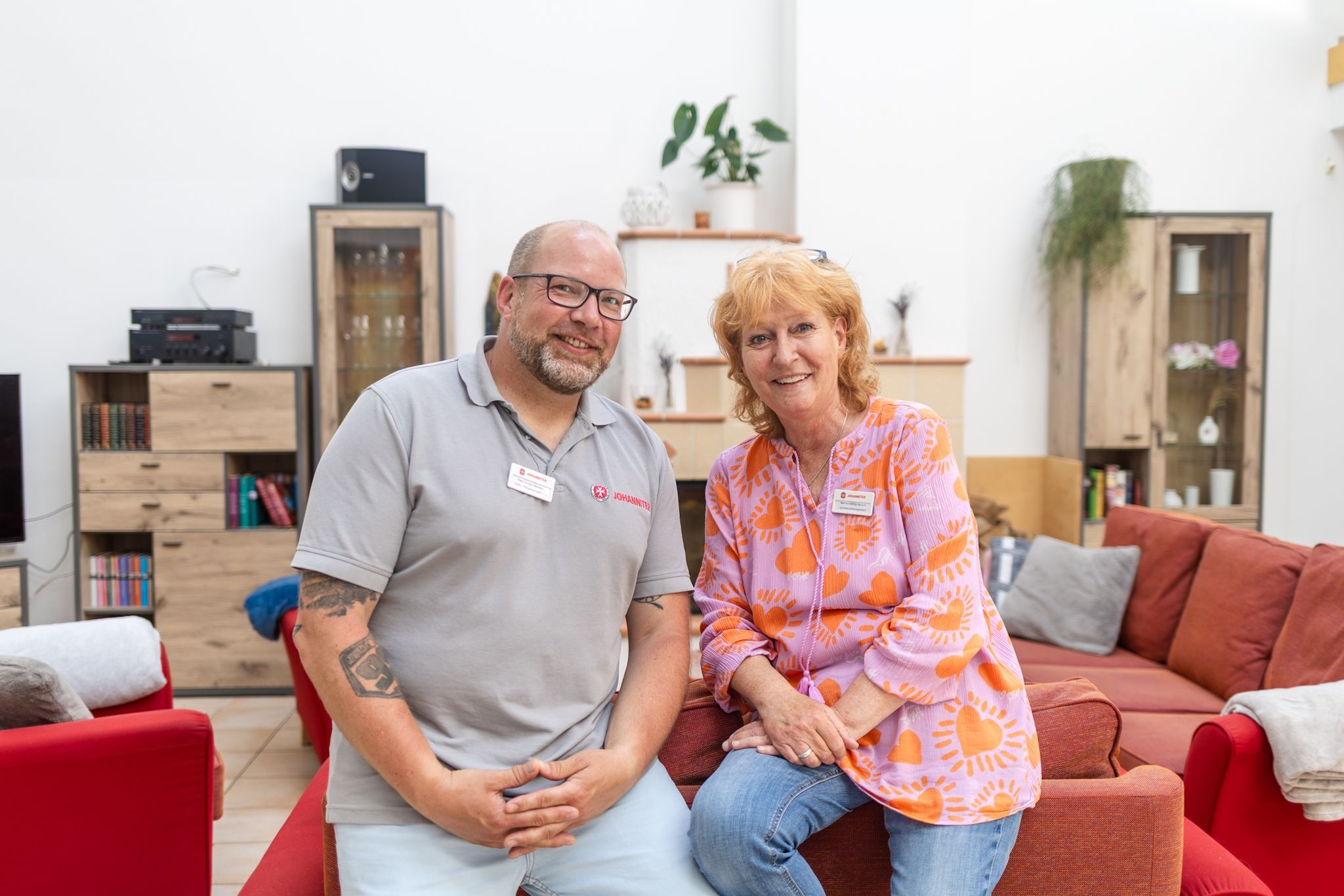 Ein Mann und eine Frau lehnen auf einer roten Couch und schauen in die Kamera.