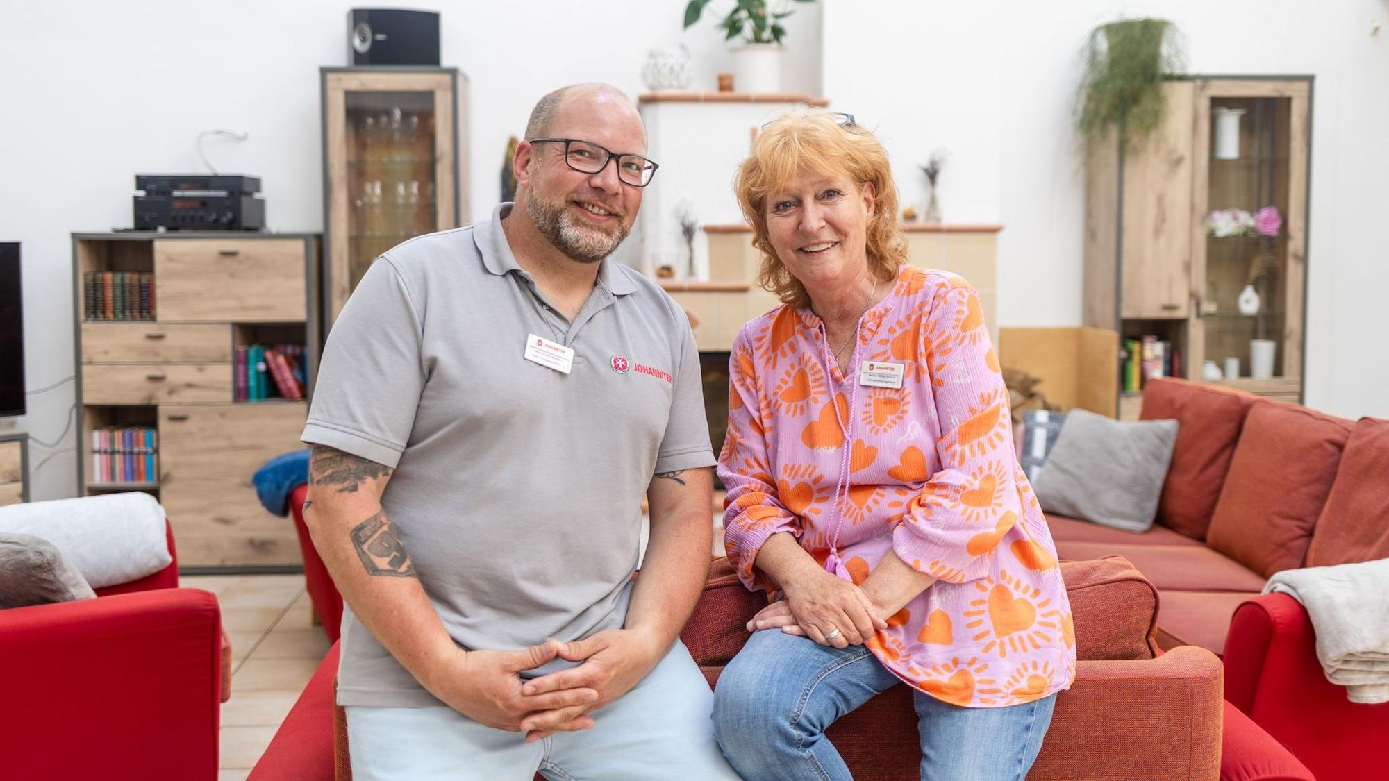 Ein Mann und eine Frau lehnen auf einer roten Couch und schauen in die Kamera.