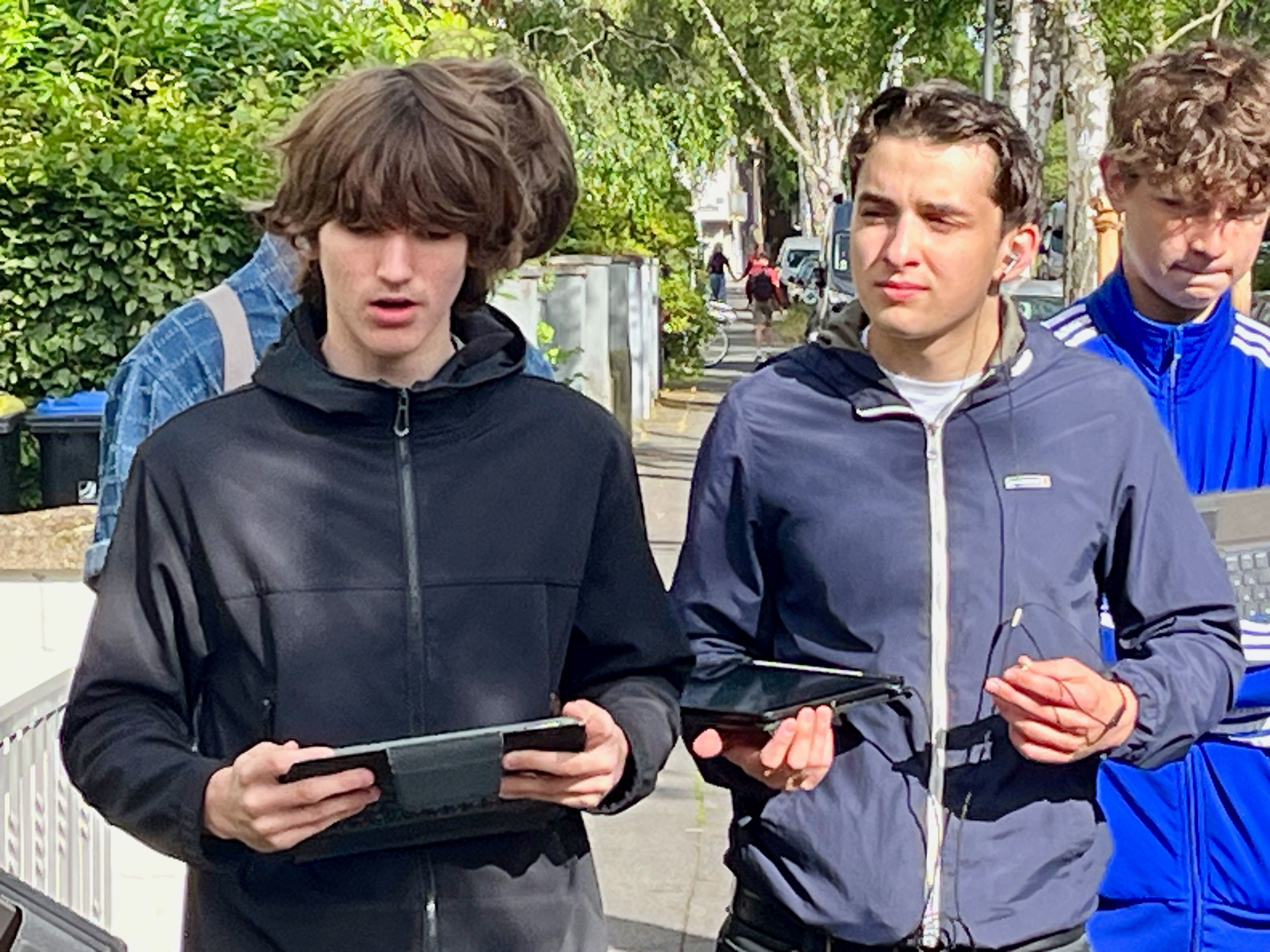 Zwei Schüler stehen auf einer Straße, zwei haben Tablets in den Händen, einer spricht.
