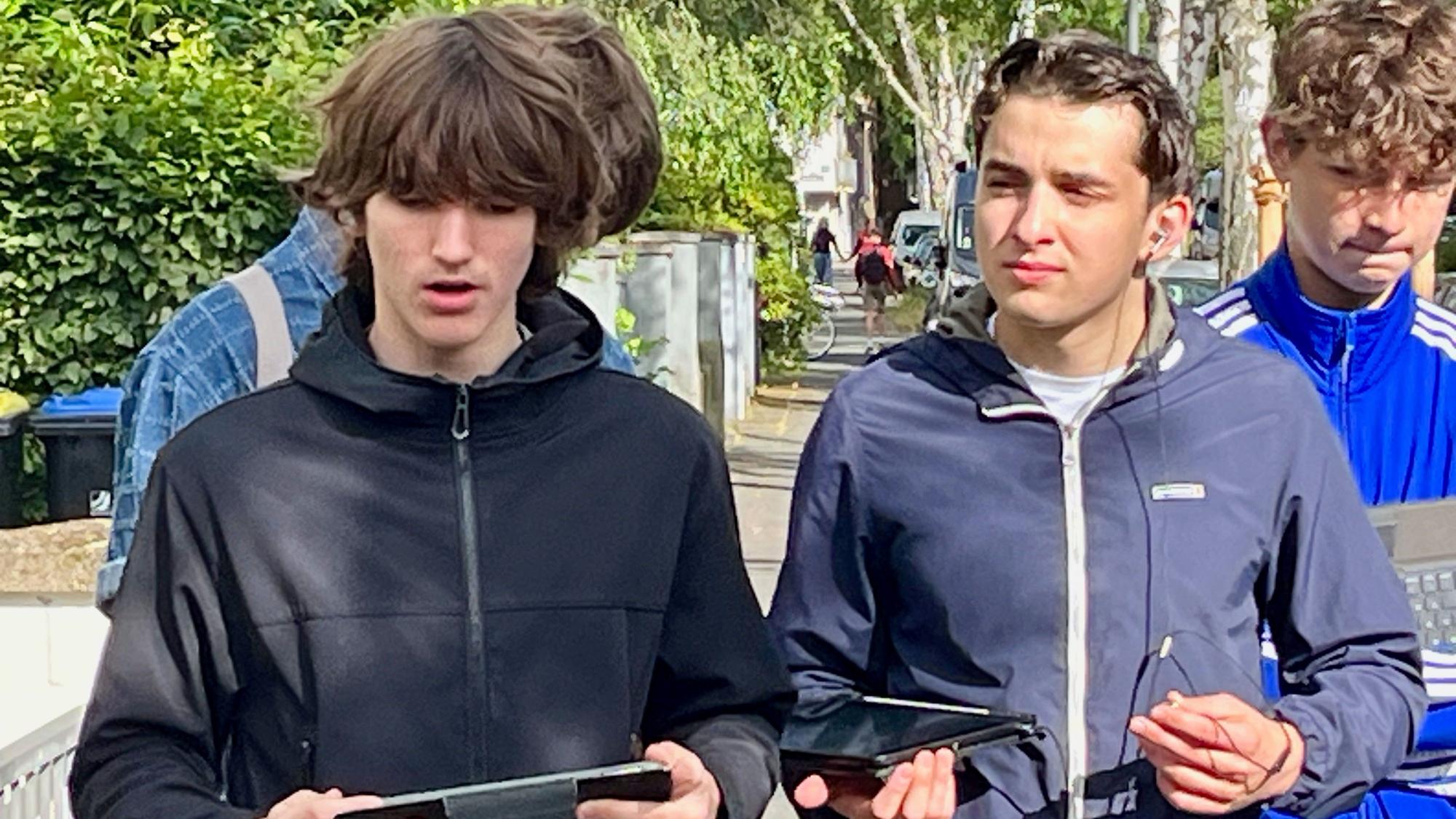 Zwei Schüler stehen auf einer Straße, zwei haben Tablets in den Händen, einer spricht.