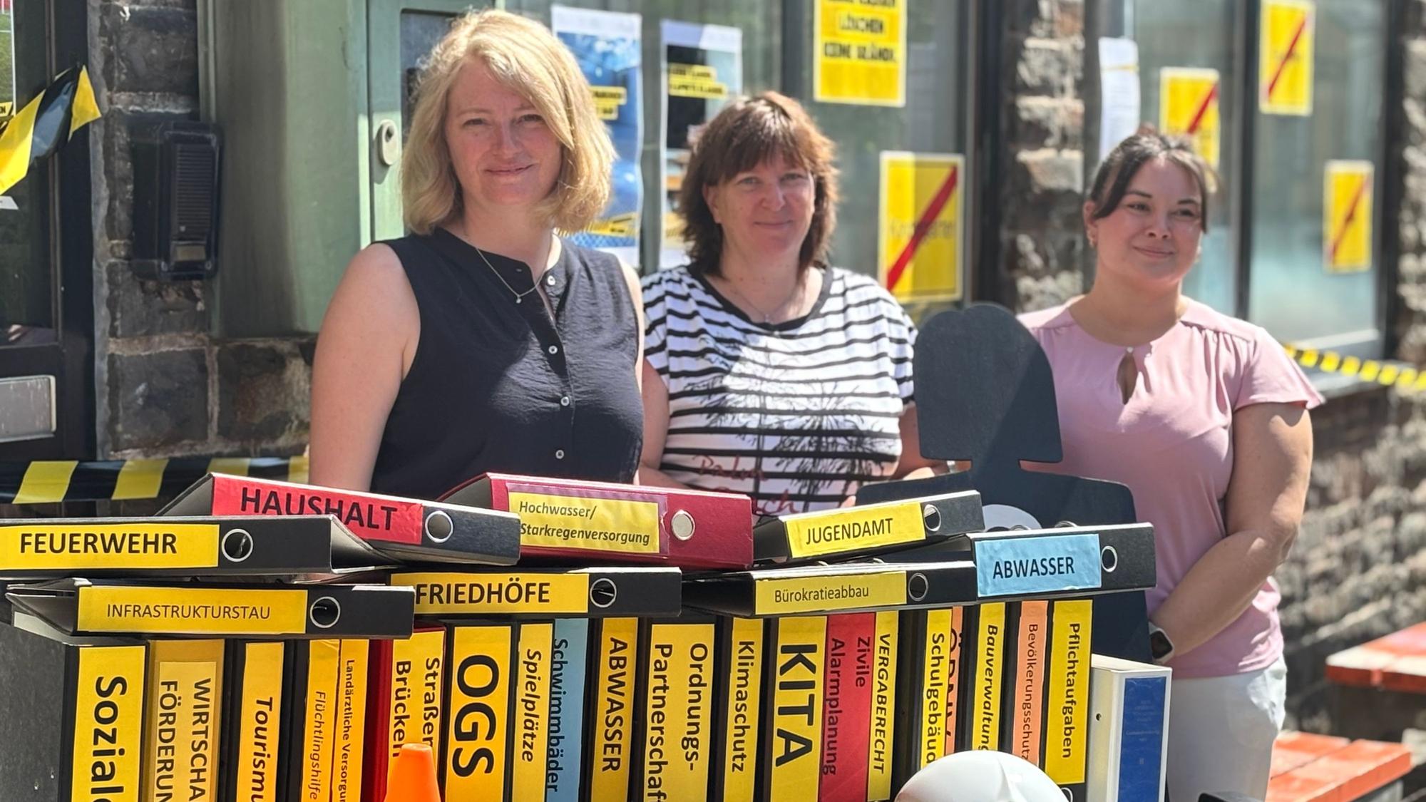 Drei Frauen stehen hinter einem Tisch auf dem Ordner mit verschiedenen Kommunalen Kostenpunkten wie „Feuerwehr, Haushalt und Jugendamt“ aufgereiht sind.