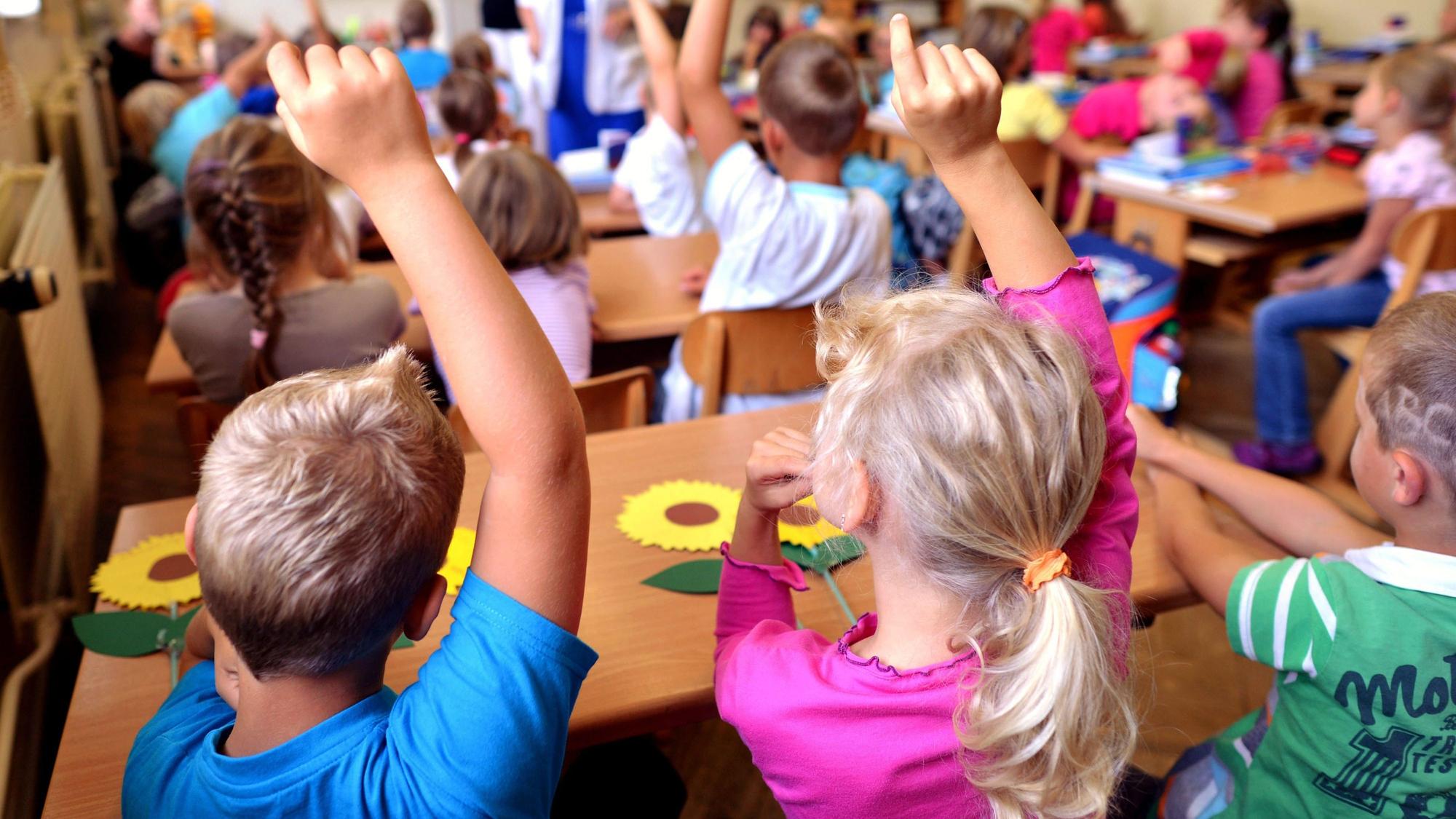 Kinder sitzen an Holztischen in einem Klassenraum.