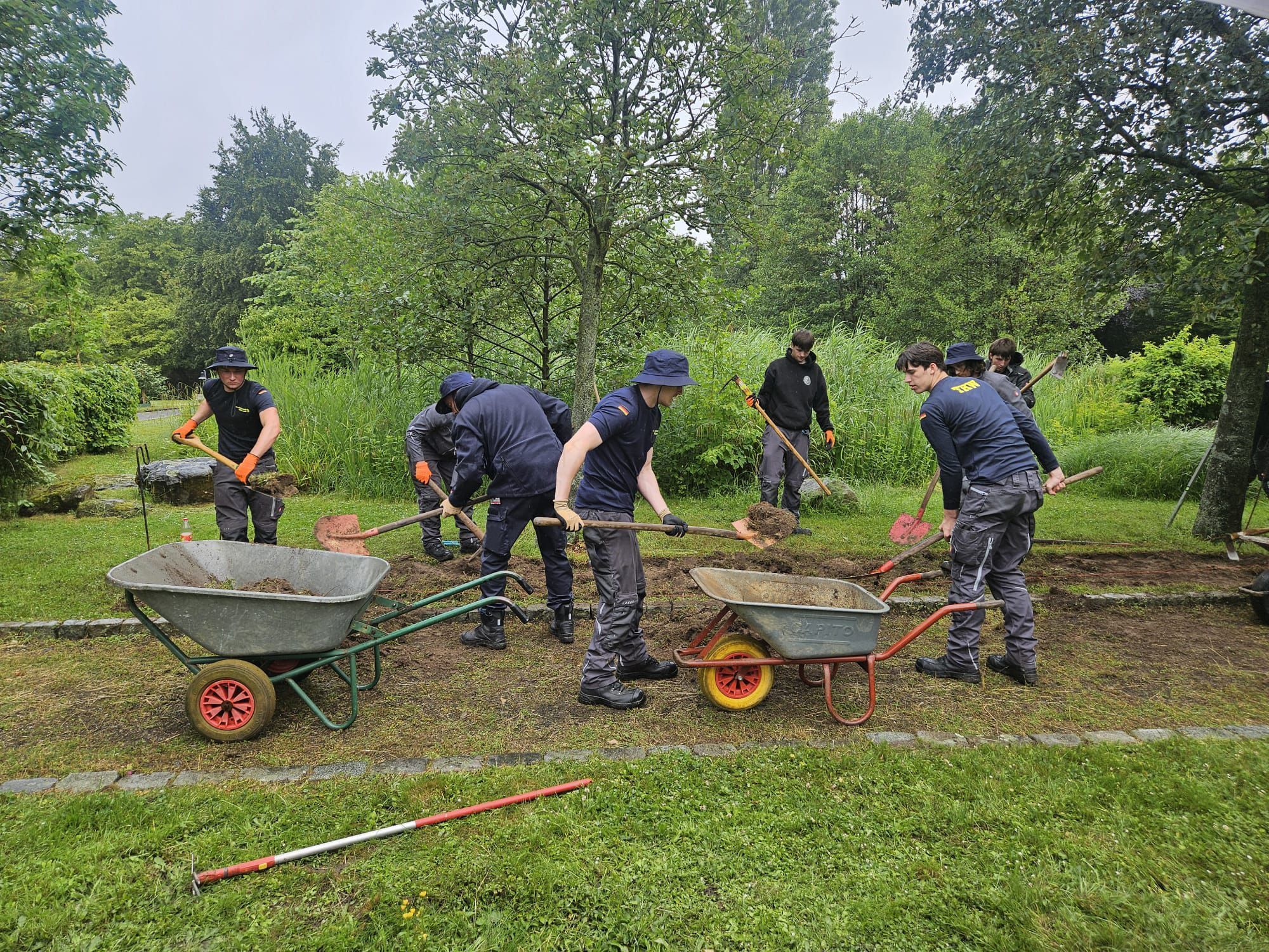 Mehrere Männer arbeiten mit Schaufeln in einer Grünanlage. Sie haben auch zwei Schubkarren dabei.
