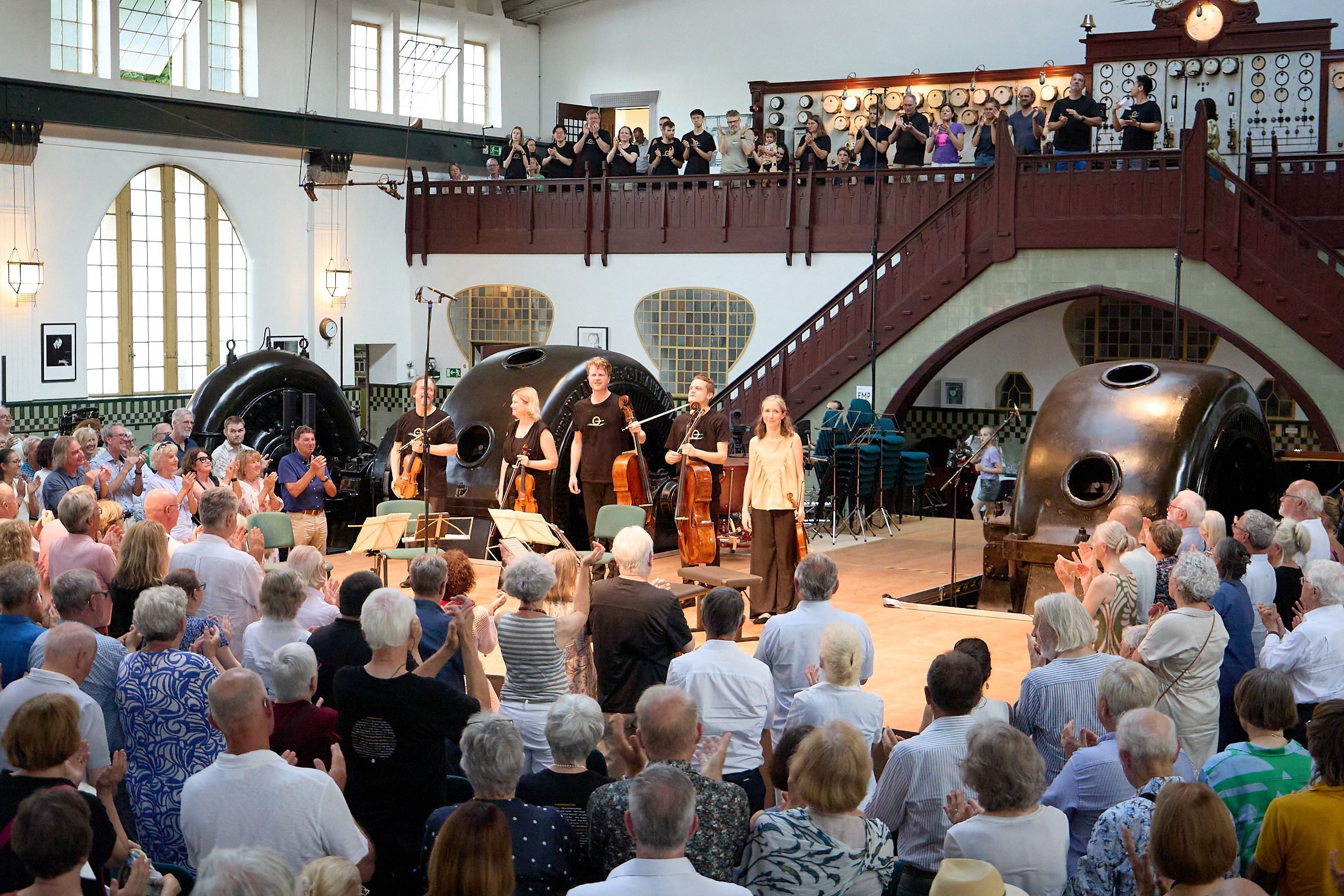 Das Foto zeigt das Publikum, das stehend applaudiert, von hinten. Auf der Bühne sind die Musiker zu sehen.