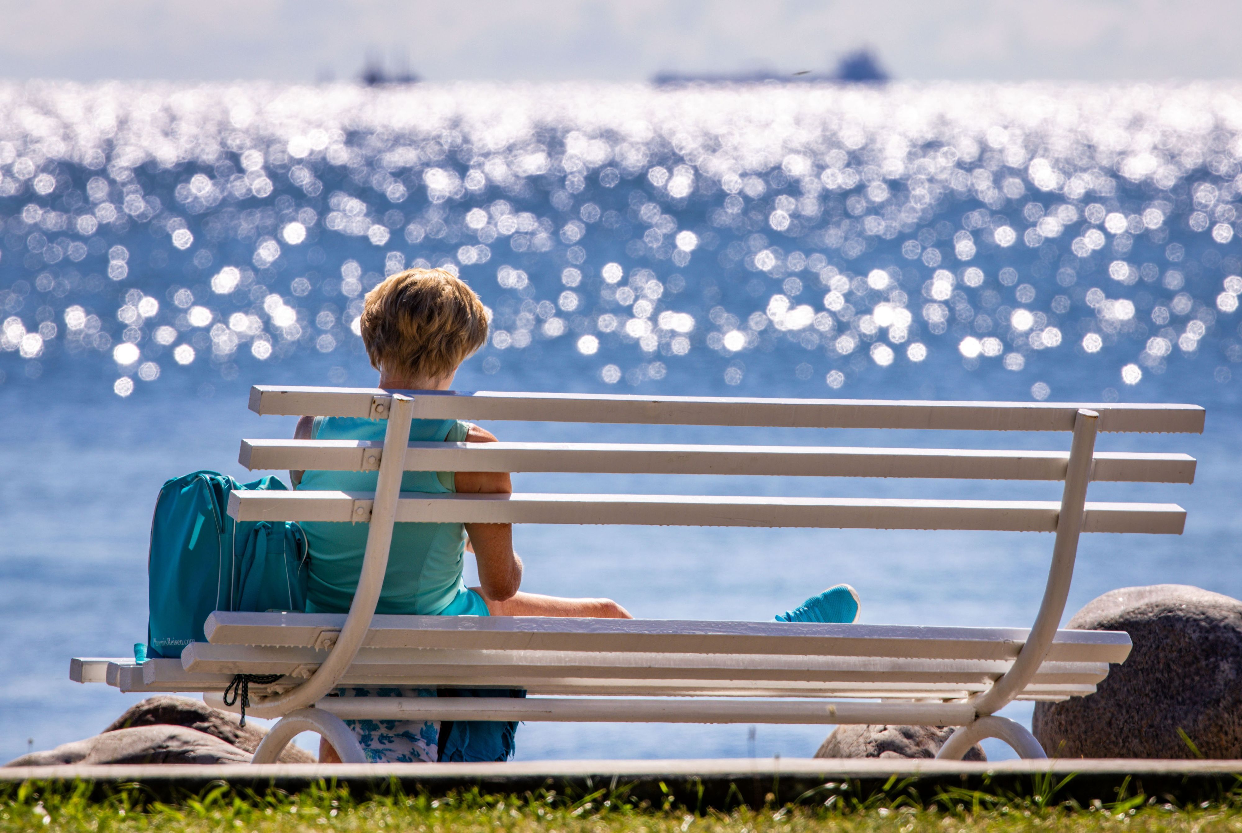 Eine Frau sitzt bei Sonne auf einer Bank am Meer und liest ein Buch. Aber Urlaub kann sich nicht jeder Mensch in Deutschland leisten. (Symbolbild)
