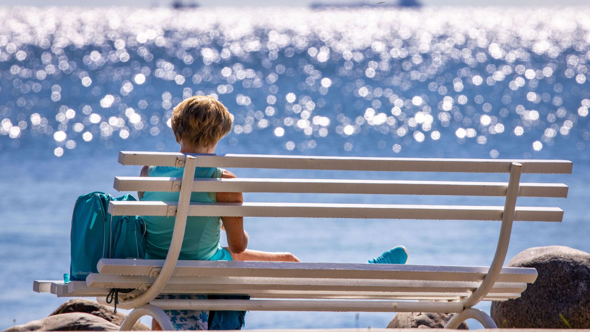 Eine Frau sitzt bei Sonne auf einer Bank am Meer und liest ein Buch. Aber Urlaub kann sich nicht jeder Mensch in Deutschland leisten. (Symbolbild)