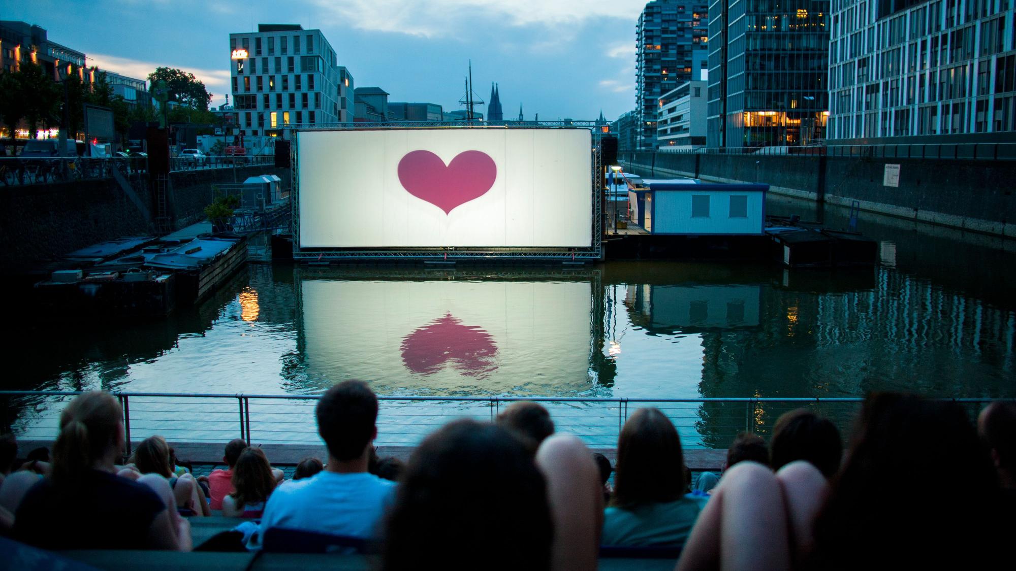 Menschen schauen sich im Open-Air-Kino am Rheinauhafen vor der Domkulisse einen Film an. (Archivbild)