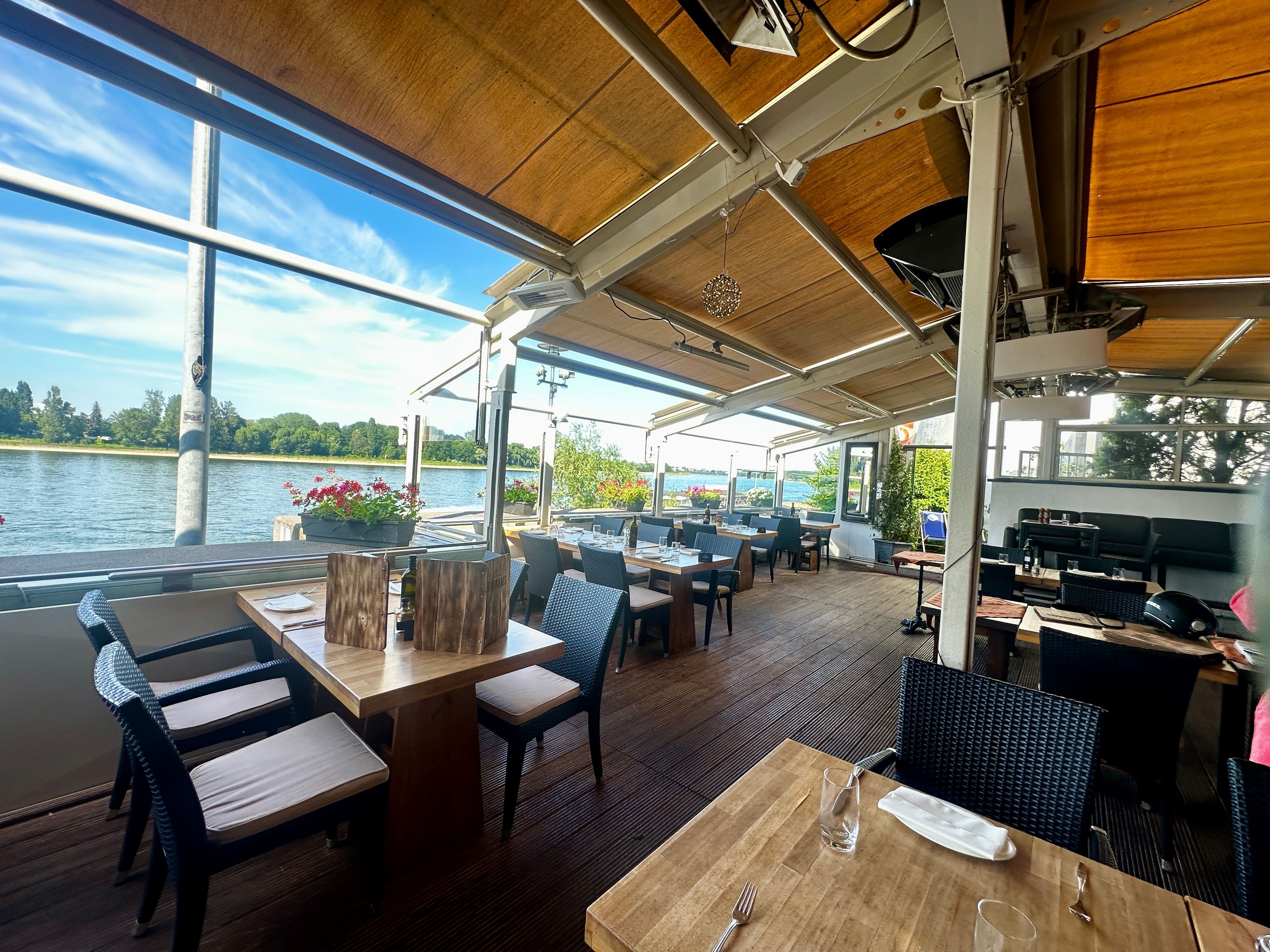 Tische stehen auf einer überdachten Terrasse, dahinter ist der Rhein zu sehen und der blaue Himmel darüber.
