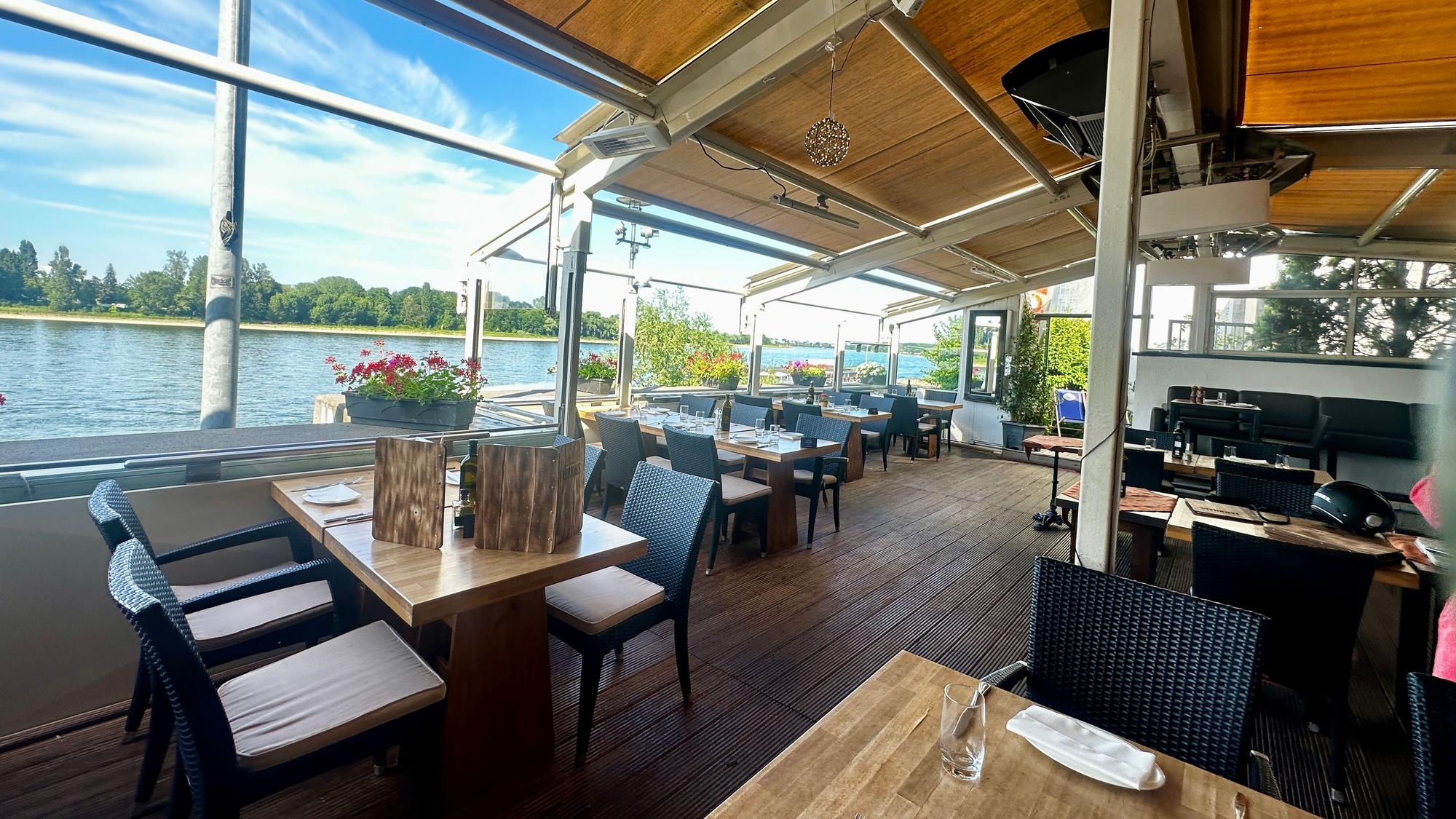 Tische stehen auf einer überdachten Terrasse, dahinter ist der Rhein zu sehen und der blaue Himmel darüber.