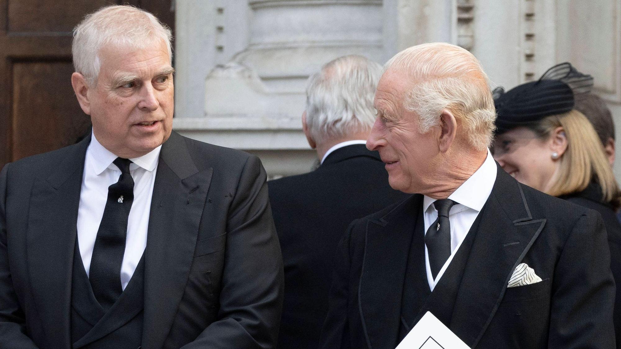 Prinz Andrew und König Charles III unterhalten sich in schwarzen Anzügen und halten Dokumente.