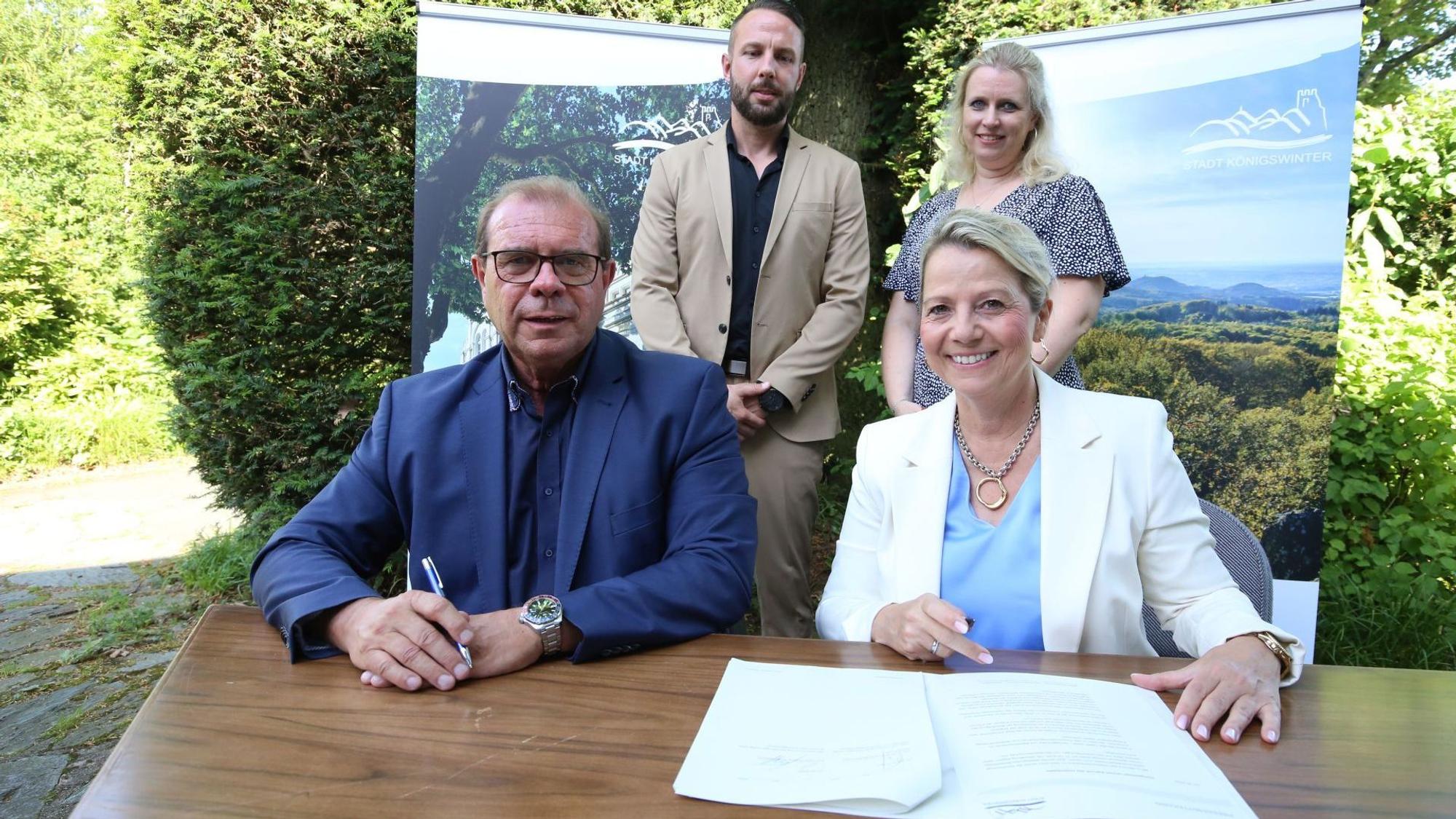 Vier Menschen; ein Mann und eine Frau sitzen an einem Tisch, auf dem ein Vertrag liegt.