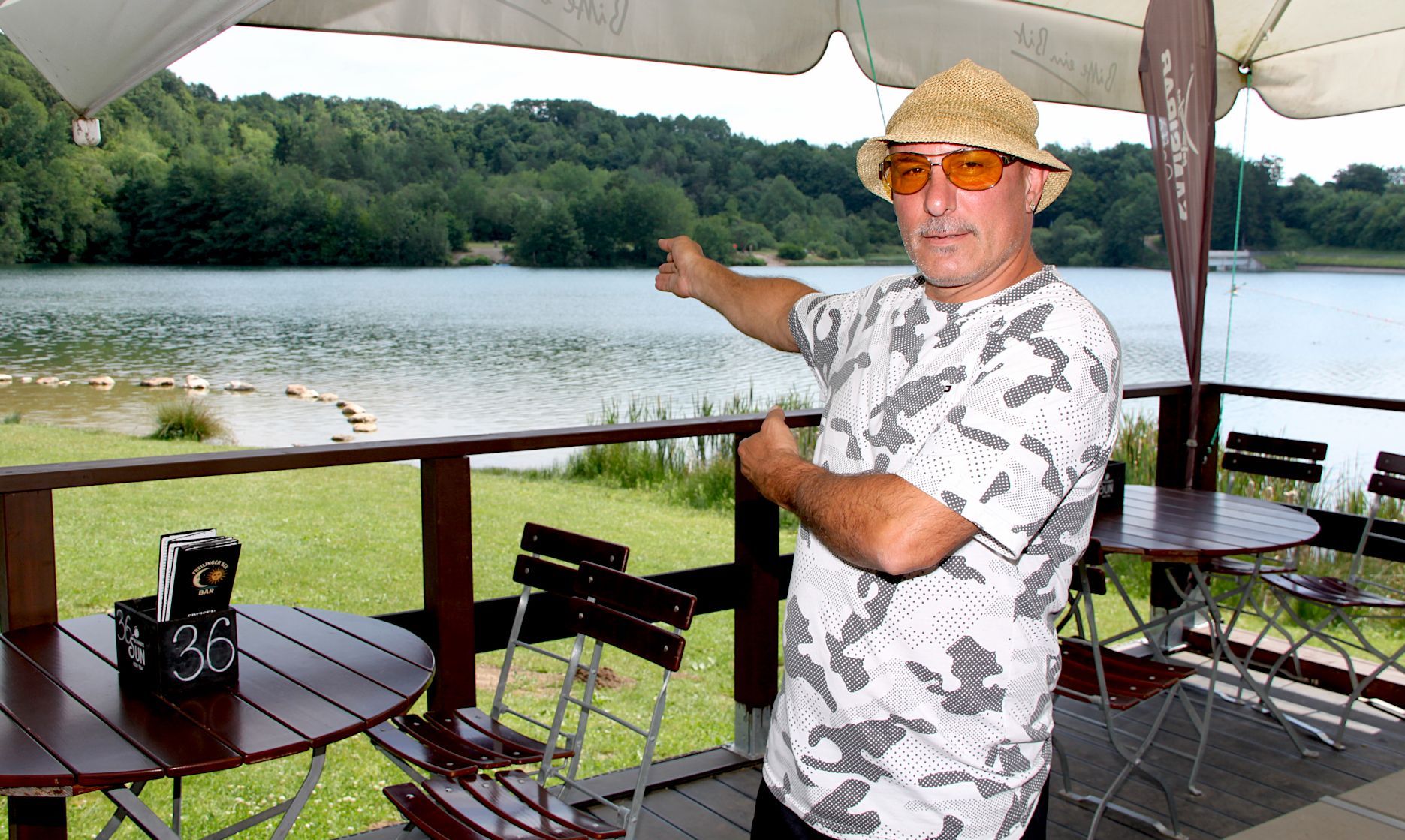 Ein Mann mit Hut steht auf einer Terrasse und zeigt auf den See.