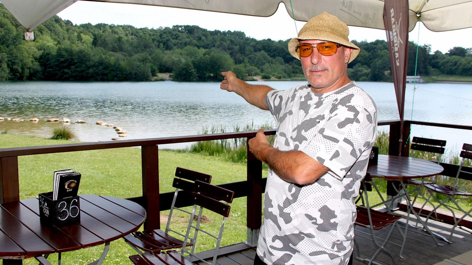 Ein Mann mit Hut steht auf einer Terrasse und zeigt auf den See.
