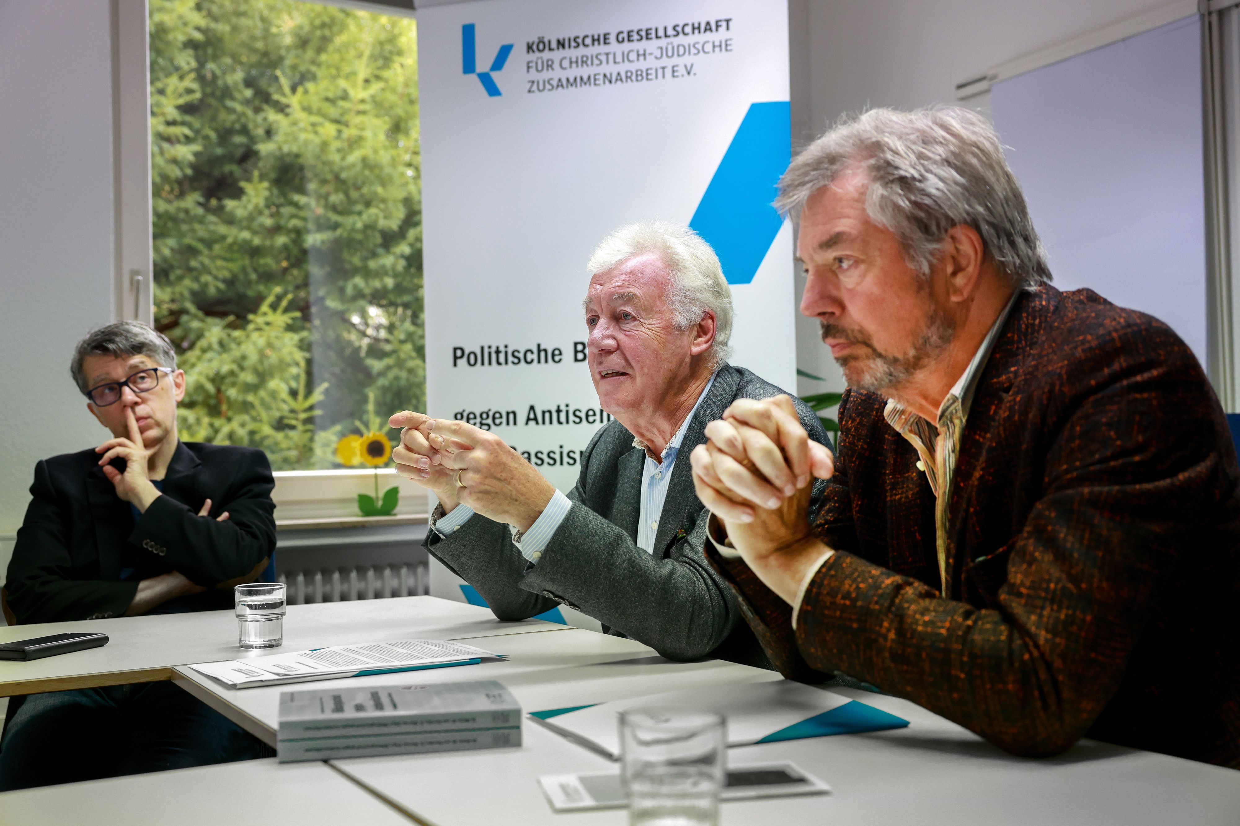 Jürgen Wilhlem sitzt bei einem Pressegsepräch an einem Tisch, im Hintergrund ist ein Aufsteller der Kölnischen Gesellschaft für Christlich-Jüdische Zusammenarbeit zu sehen.