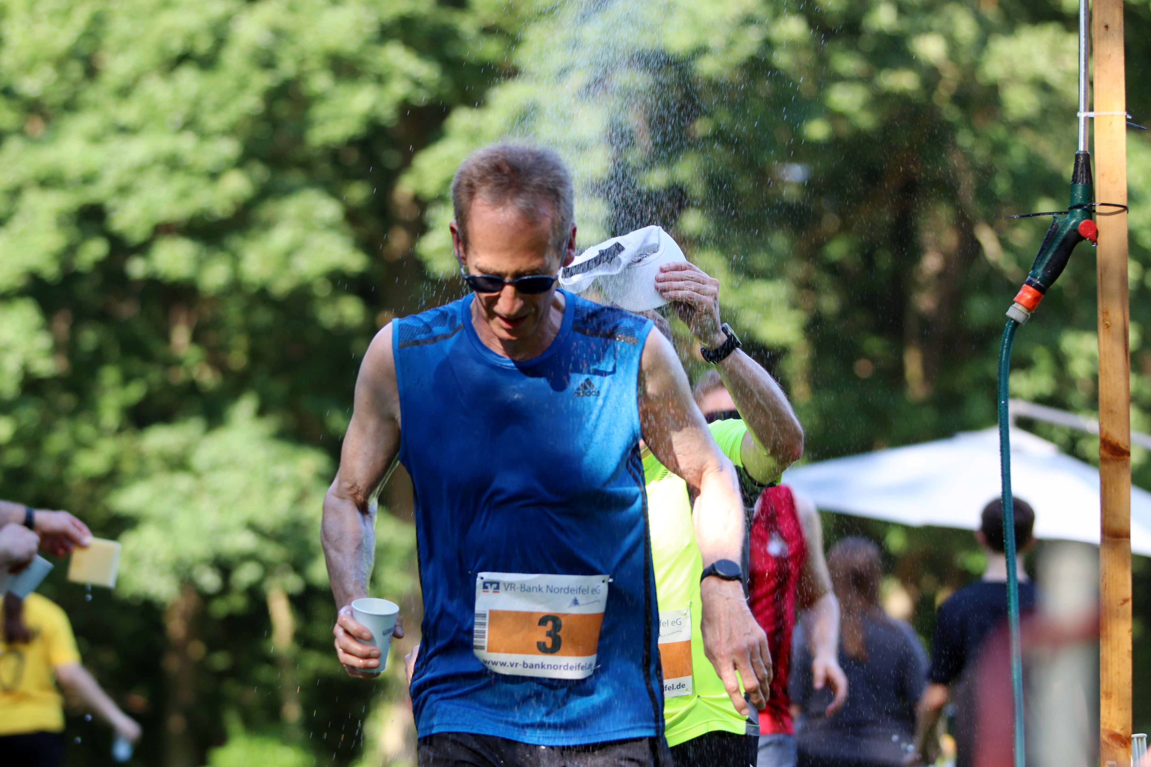 Detlef Seif läuft, mit einem Becher Wasser in der Hand, durch die Streckendusche.