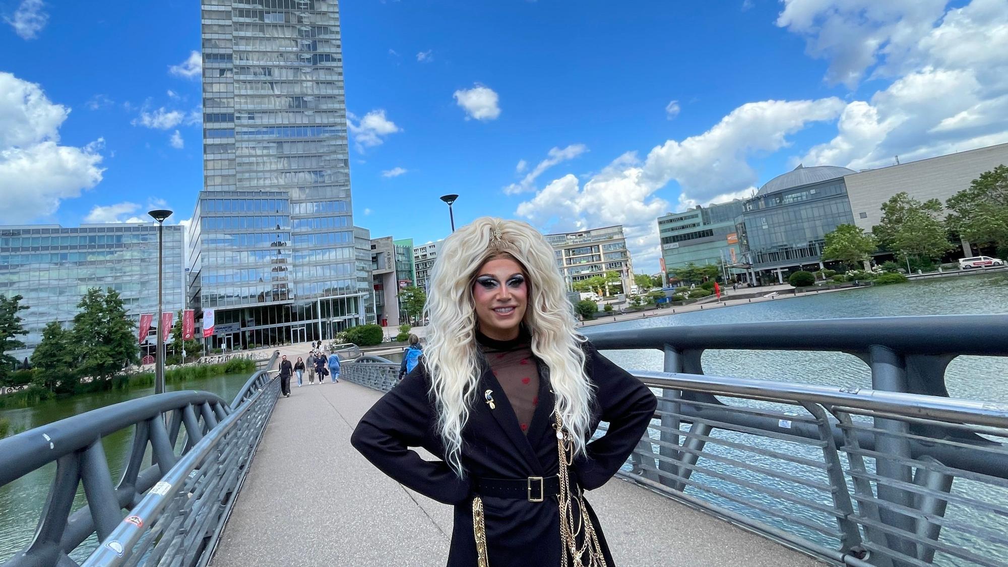 Eine Drag Queen mit blonder langer Perücke und schwarzem Mantel steht auf einer Brücke, im Hintergrund ist der Mediapark-Turm zu sehen.