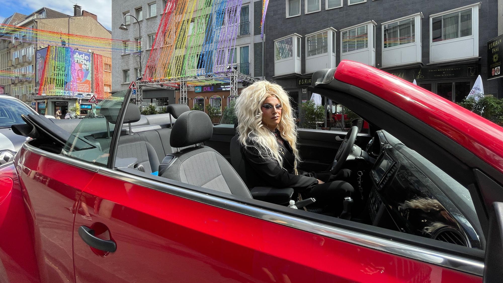 Ein als Frau mit langem blonden Haar verkleideter Mann sitzt in einem roten Cabrio. Im Hintergrund sind Fassaden mit Regenbogen-Deko zu sehen.