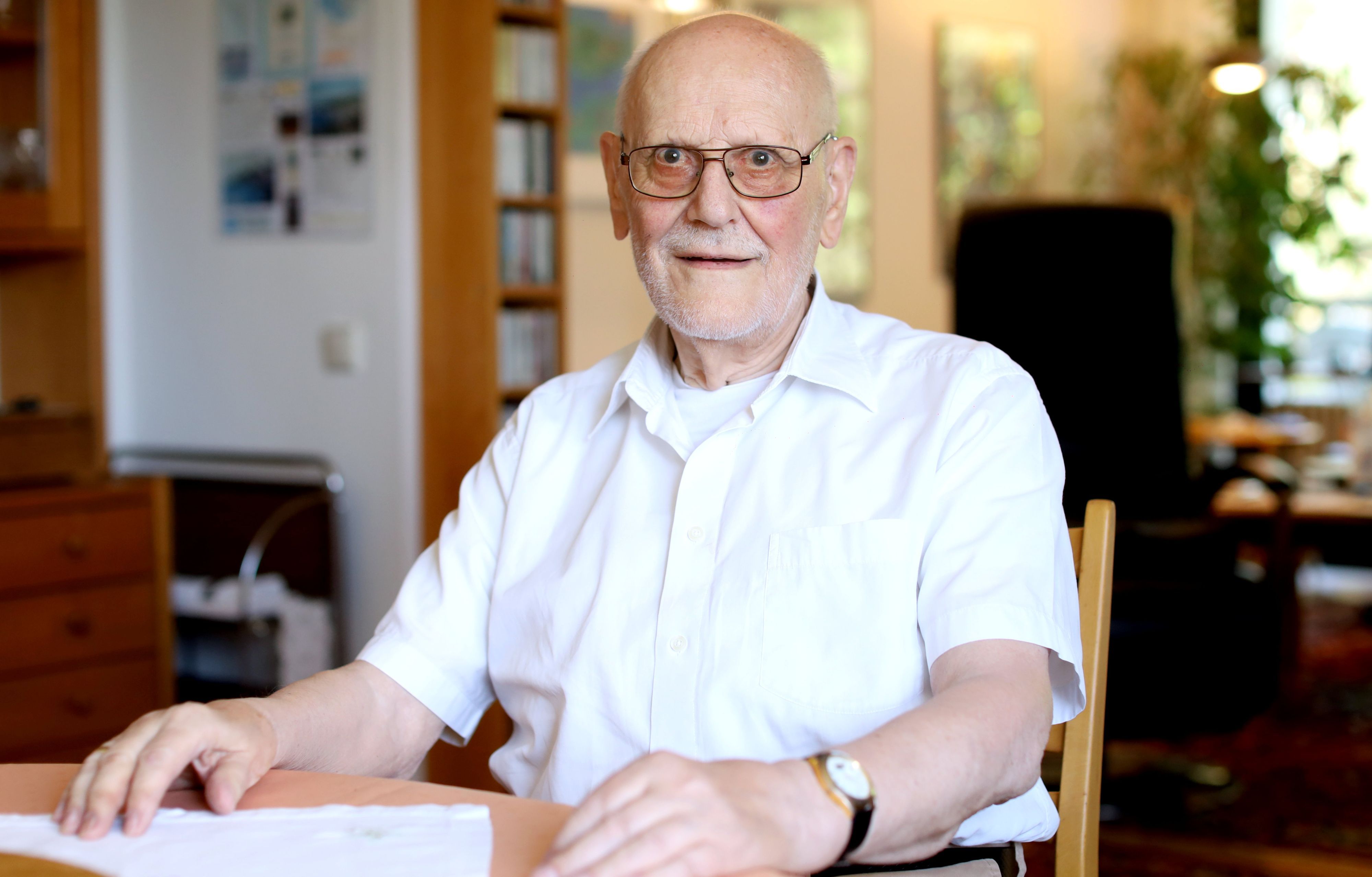 Werner Löwenstein sitzt an einem Tisch in seiner Wohnung und lächelt in die Kamera.