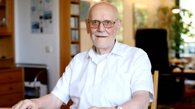 Werner Löwenstein sitzt an einem Tisch in seiner Wohnung und lächelt in die Kamera.