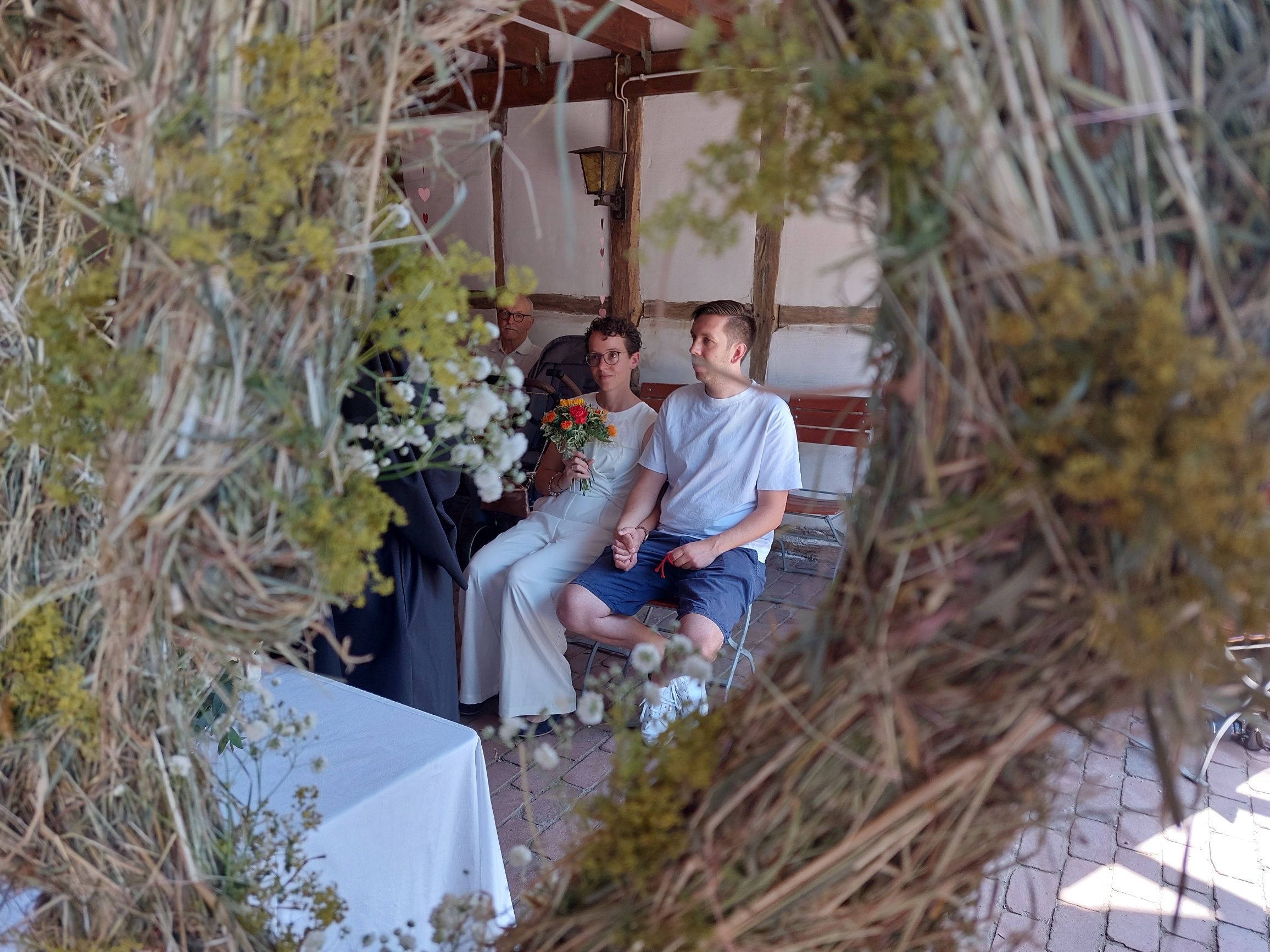 Ein Hochzeitspaar bei der Trauung: Die Braut trägt einen schicken weißen Hosenanzug, der Bräutigam Shorts und T-Shirt.