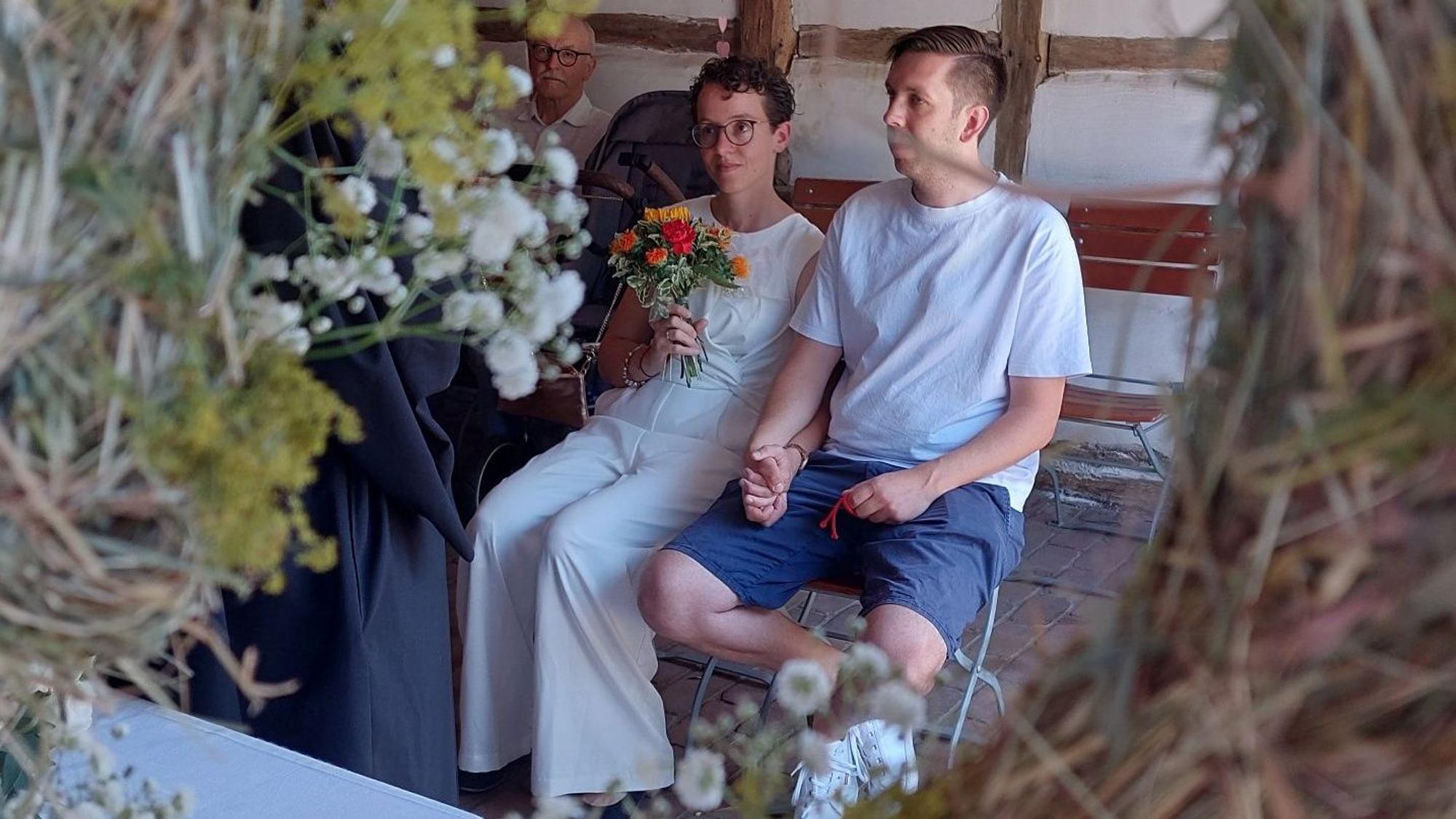 Ein Hochzeitspaar bei der Trauung: Die Braut trägt einen schicken weißen Hosenanzug, der Bräutigam Shorts und T-Shirt.