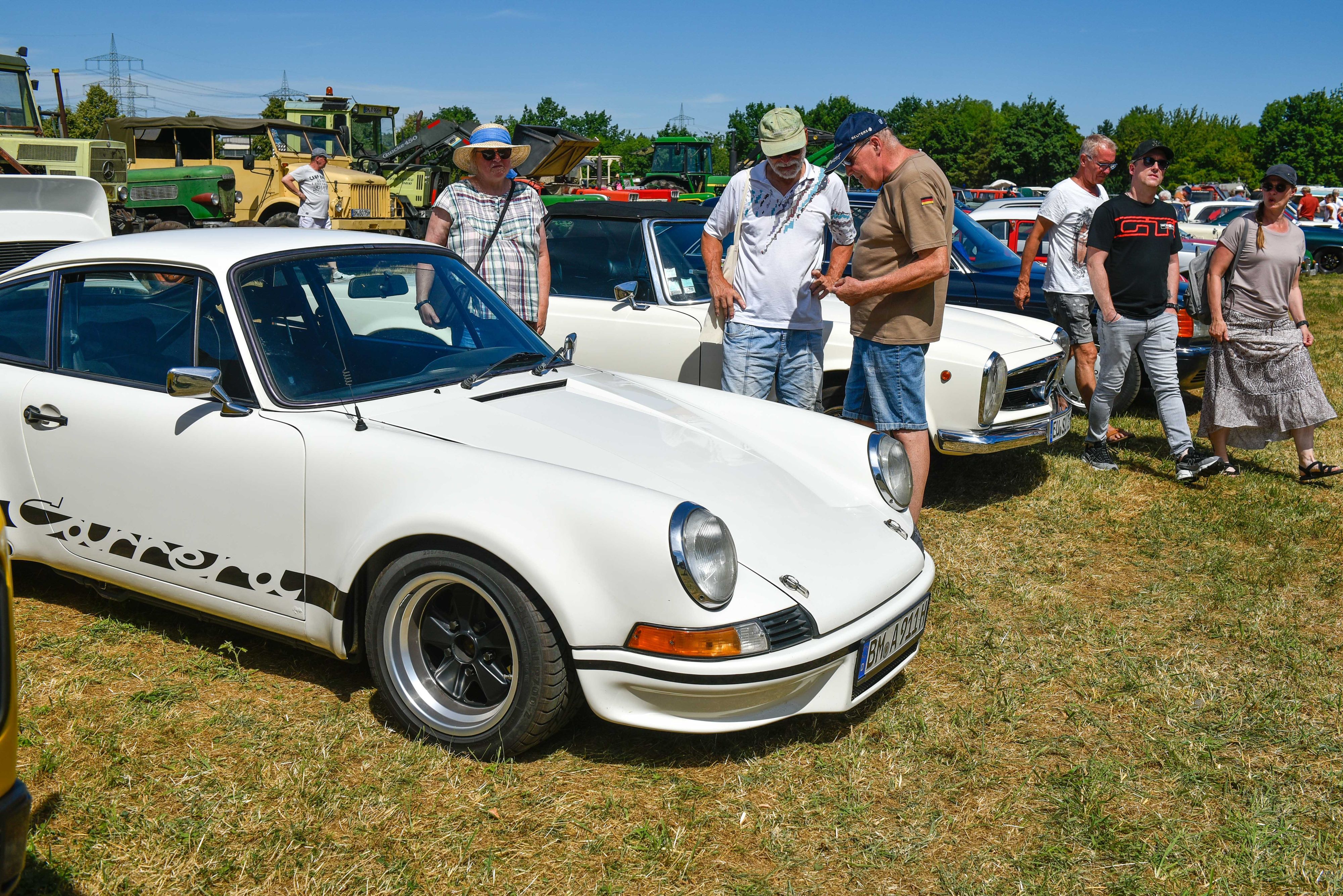 Zu sehen sind mehrere Menschen, die auf einer Wiese Porsche- und Mercedes-Oldtimer in Augenschein nehmen.