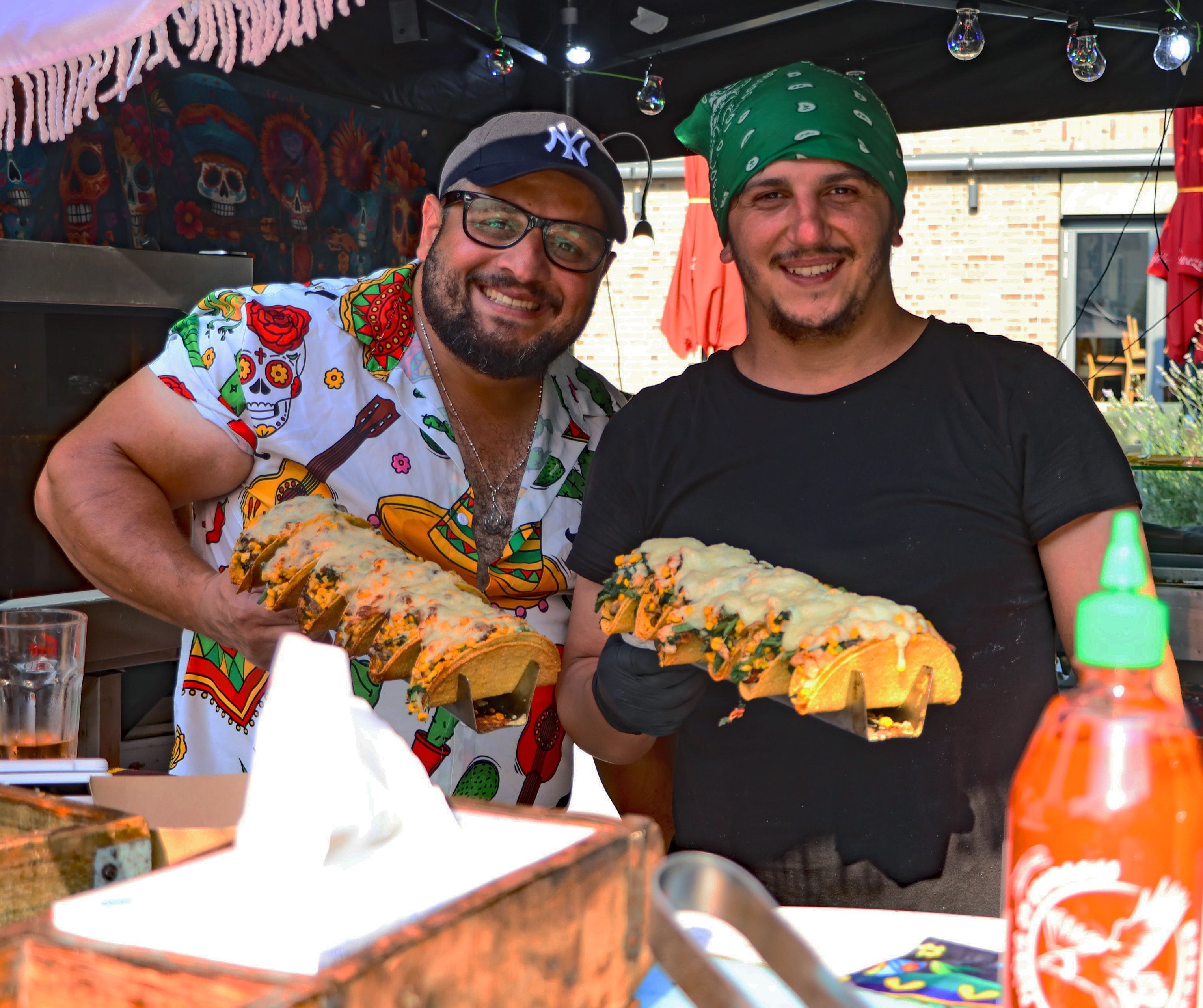 Zwei Verkäufer hinter einem Stand halten Tacos in den Händen und lächeln.