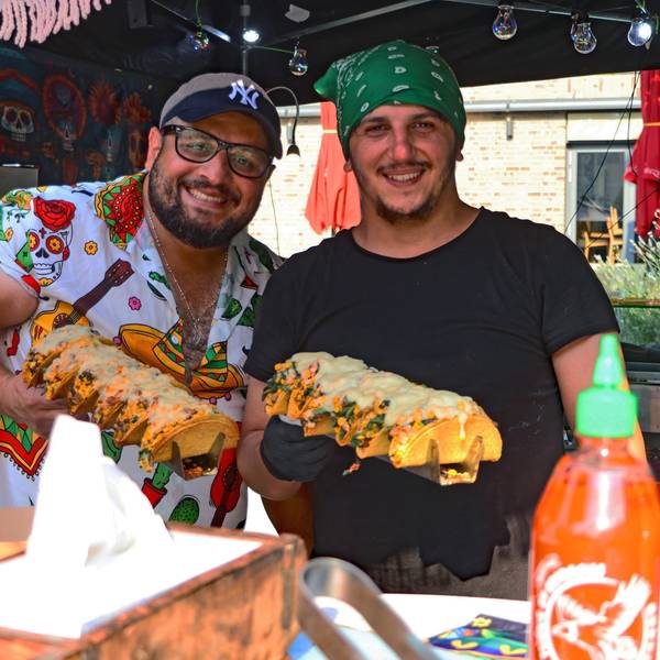 Zwei Verkäufer hinter einem Stand halten Tacos in den Händen und lächeln.