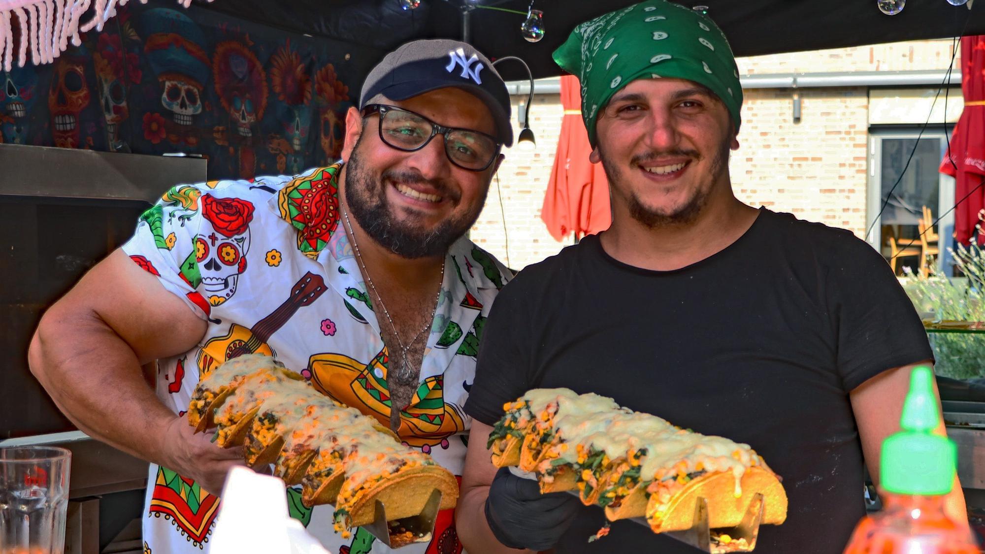 Zwei Verkäufer hinter einem Stand halten Tacos in den Händen und lächeln.