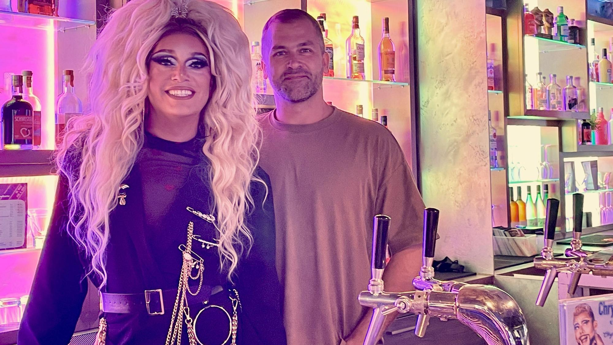 Eine Drag Queen mit langer blonder Perücke steht hinter dem Tresen einer Bar neben einem Mann im braunen Pulli mit Dreitagebart.