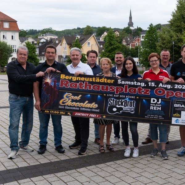 Bei einem Gruppenfoto halten mehrere Menschen ein Banner, auf dem Werbung für das Rathausplatz-Open-Air in Bergneustadt am 15. August zu lesen ist.