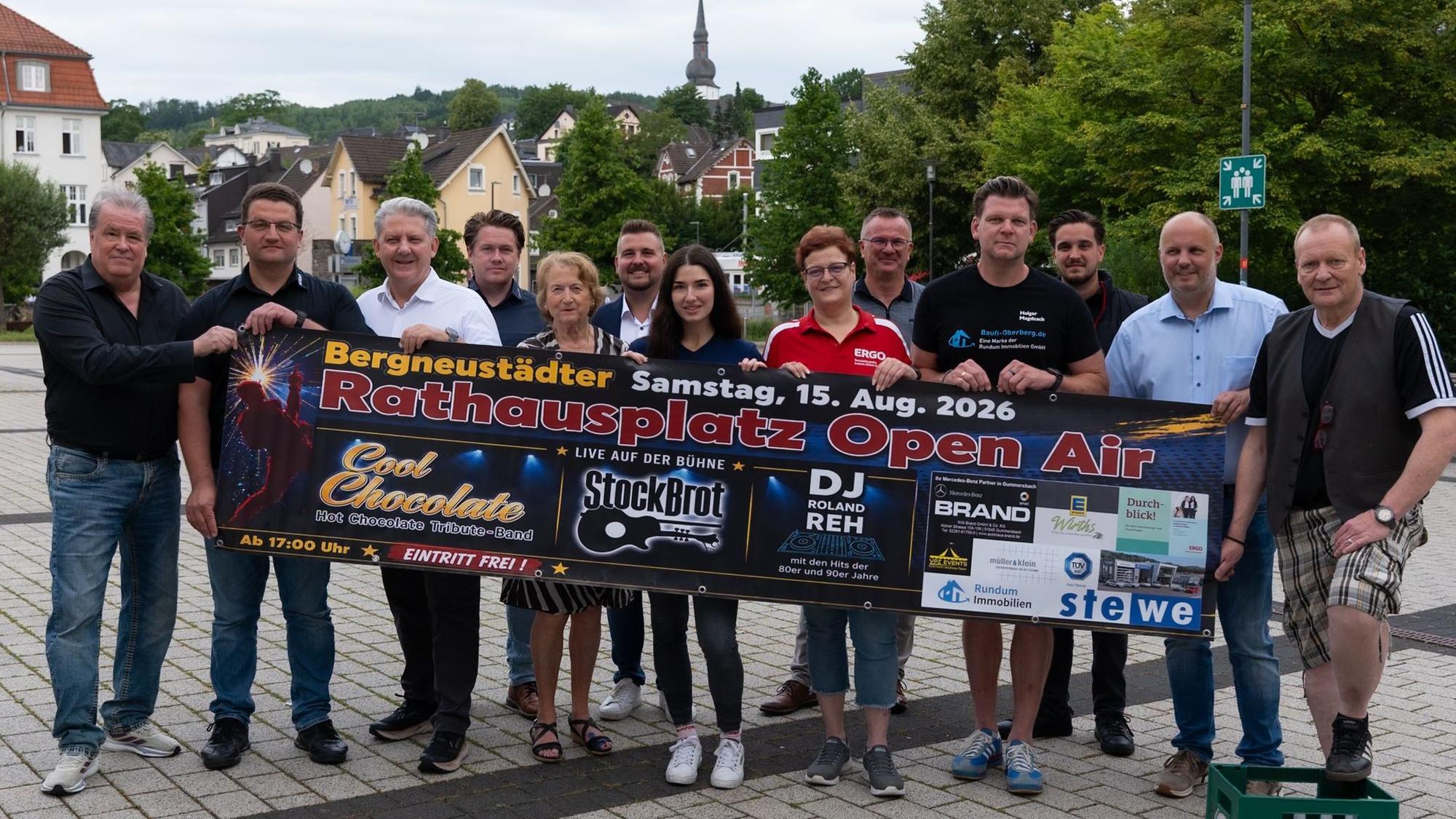 Bei einem Gruppenfoto halten mehrere Menschen ein Banner, auf dem Werbung für das Rathausplatz-Open-Air in Bergneustadt am 15. August zu lesen ist.
