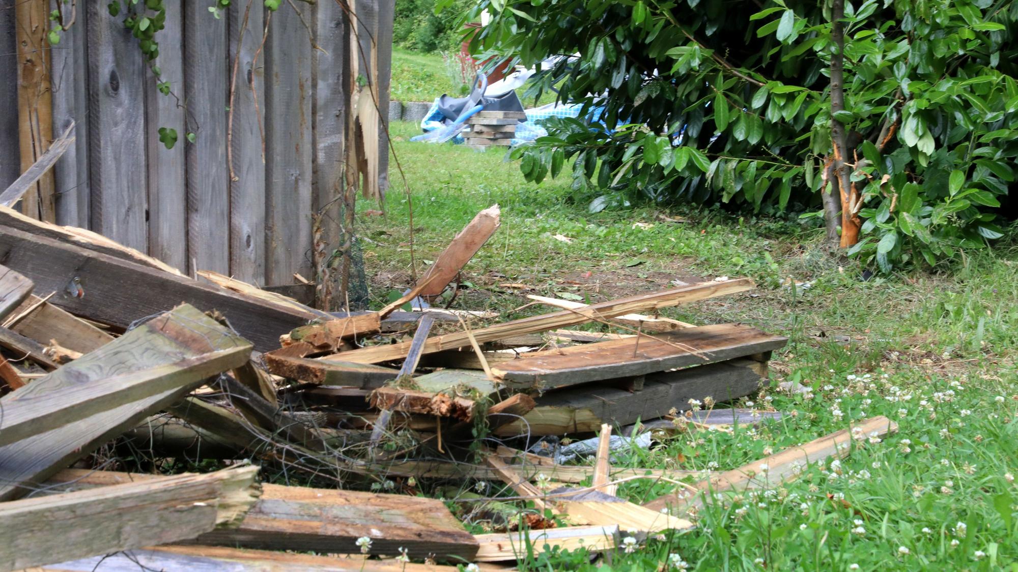 Teile eines zerstörten Bretterzauns liegen auf einer Wiese, im Hintergrund sind ein Kirschlorbeerbaum und Teile eines zerstörten Gartenpools zu sehen.