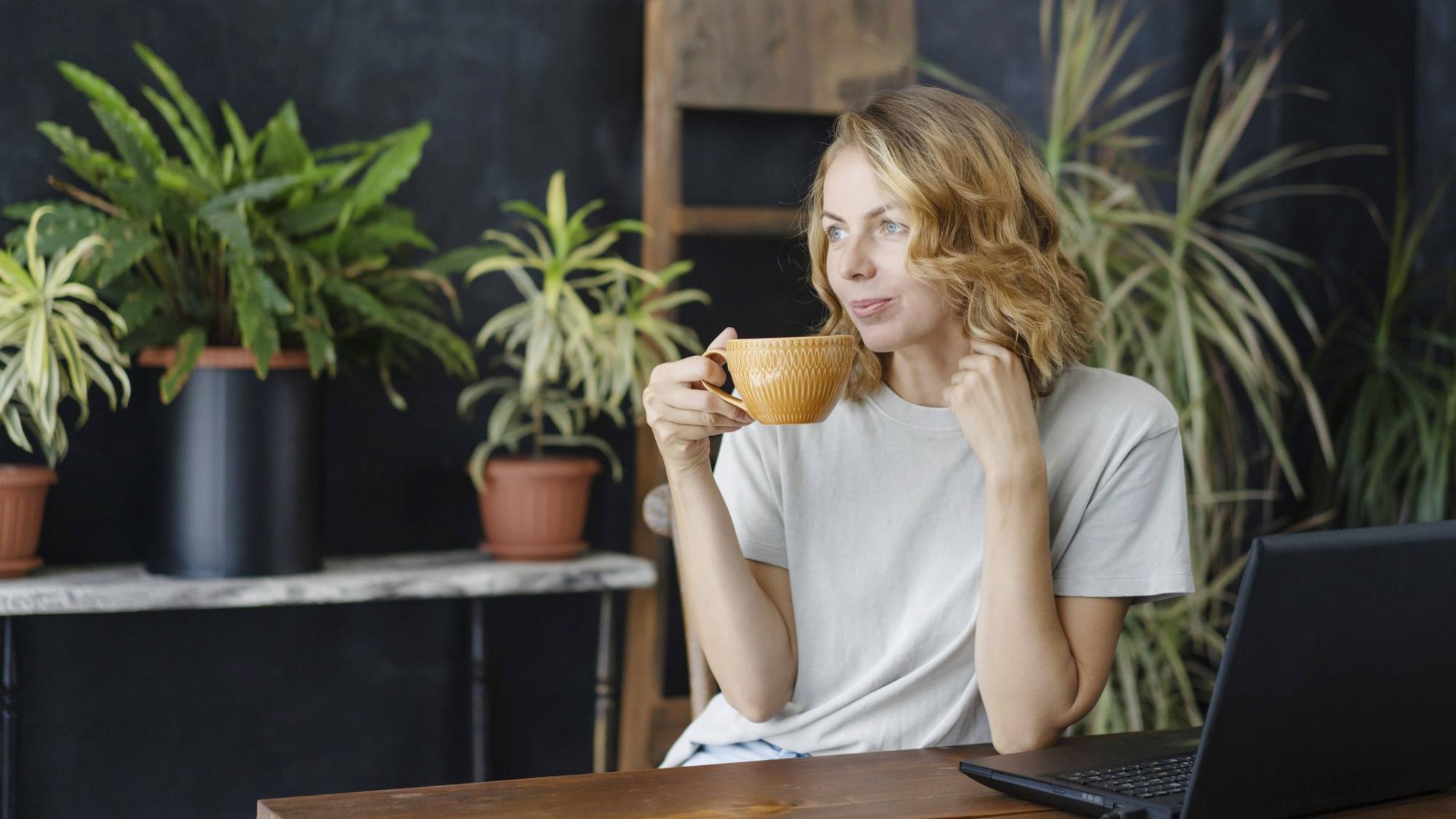 Eine Frau trinkt Kaffee und genießt an ihrem Arbeitstisch mit einem Laptop in einem gemütlichen Heimbüro mit tropischen Pflanzen im Hintergrund.