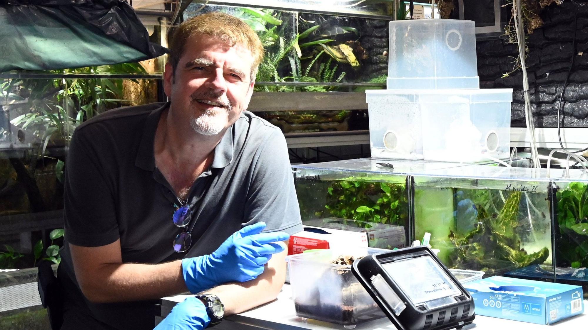 Ein Mann mit blauen Handschuhen lehnt auf einem Tisch. Im Hintergrund viele Aquarien.