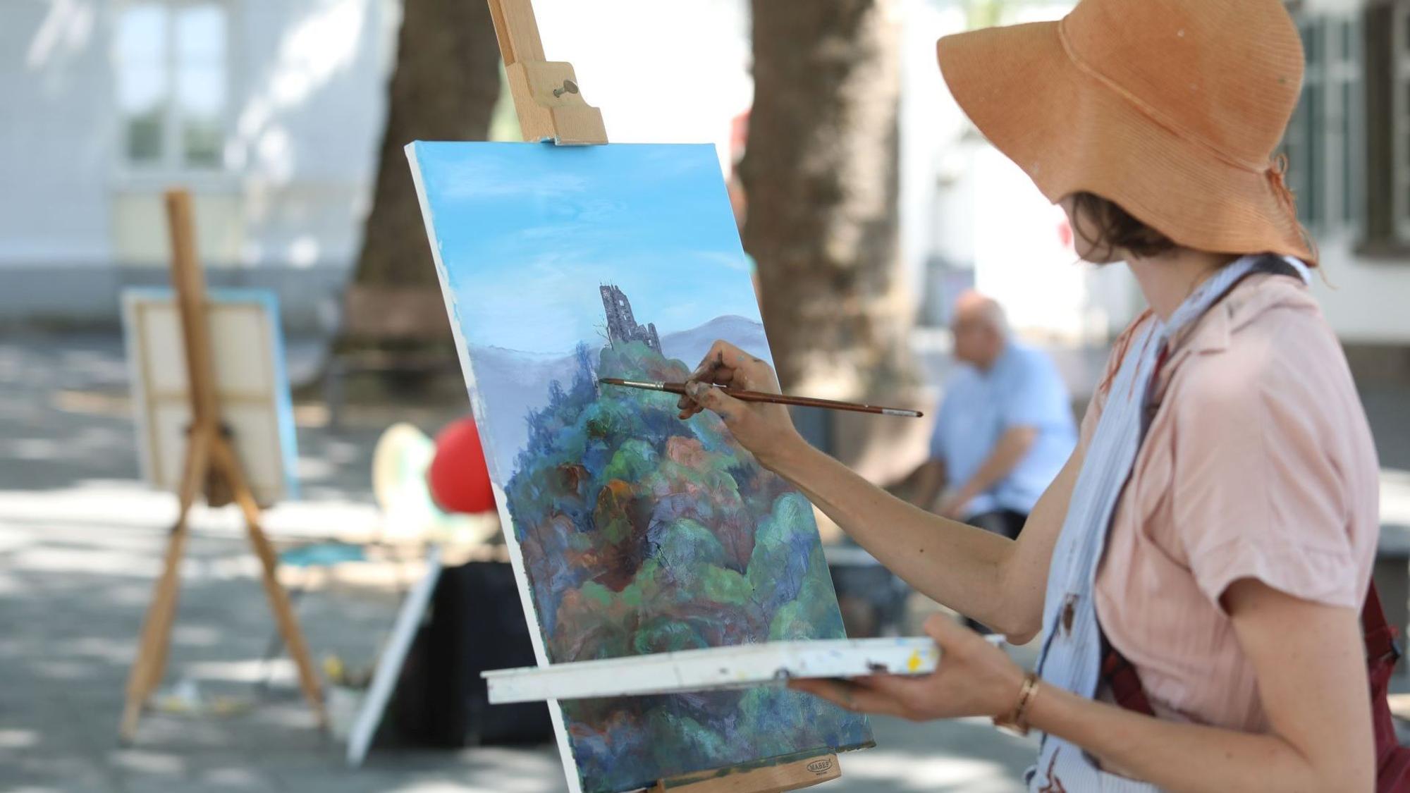 Eine Frau mit Sonnenhut malt ein Bild, das auf einer Staffelei steht und das den Drachenfels zeigt.