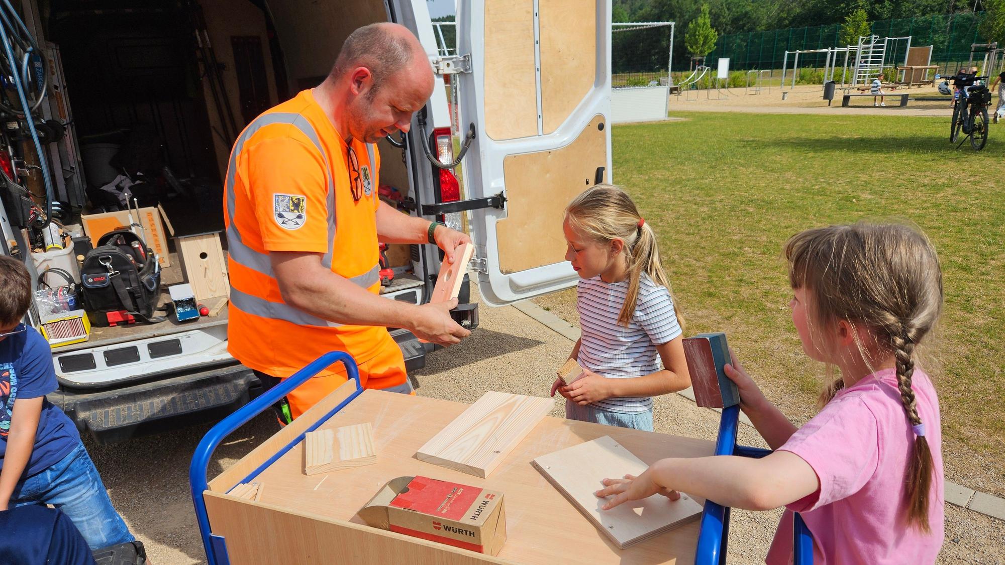 Ein Mitarbeiter des Bauhofs und zwei Mädchen arbeiten mit Holzteilen.