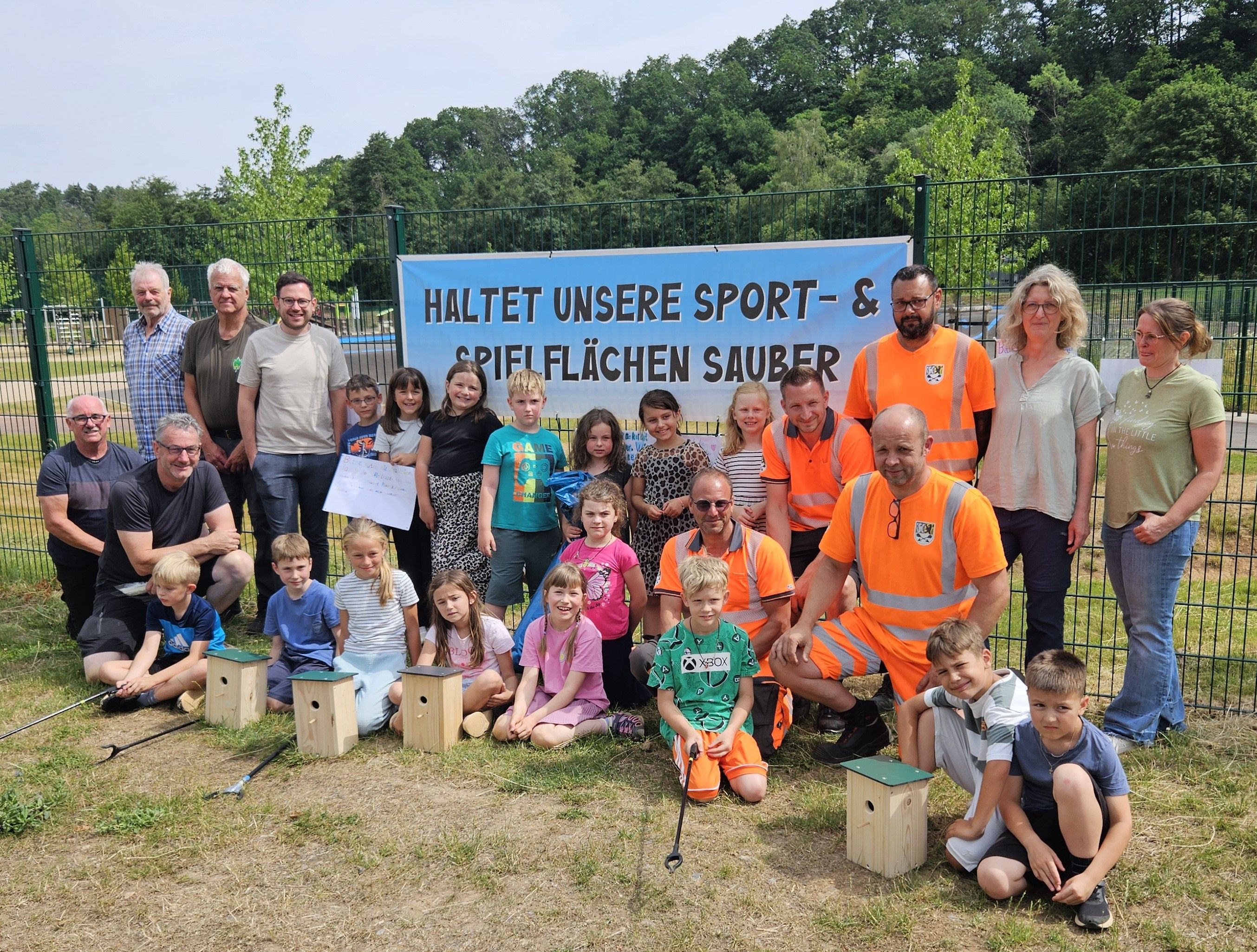 Die Helfer und die Kinder stehen und knien vor einem Banner mit der Aufschrift „Haltet unsere Sport- und Spielflächen sauber“.