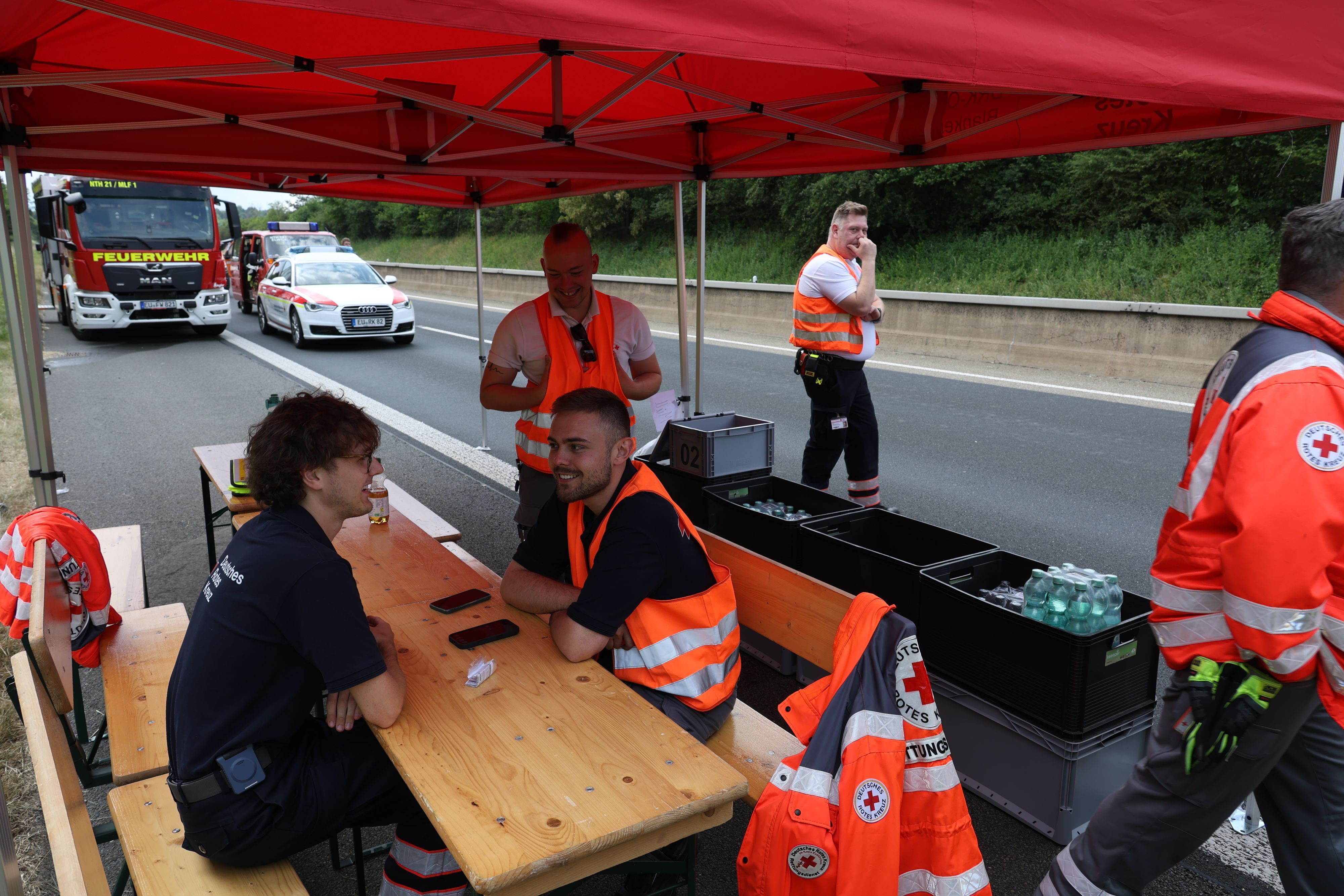 DRK-Helfer sitzen im Zelt auf der Autobahn vor Kisten mit Waserflaschen, im Hintergrund sind Einsatzfahrzeuge zu sehen.