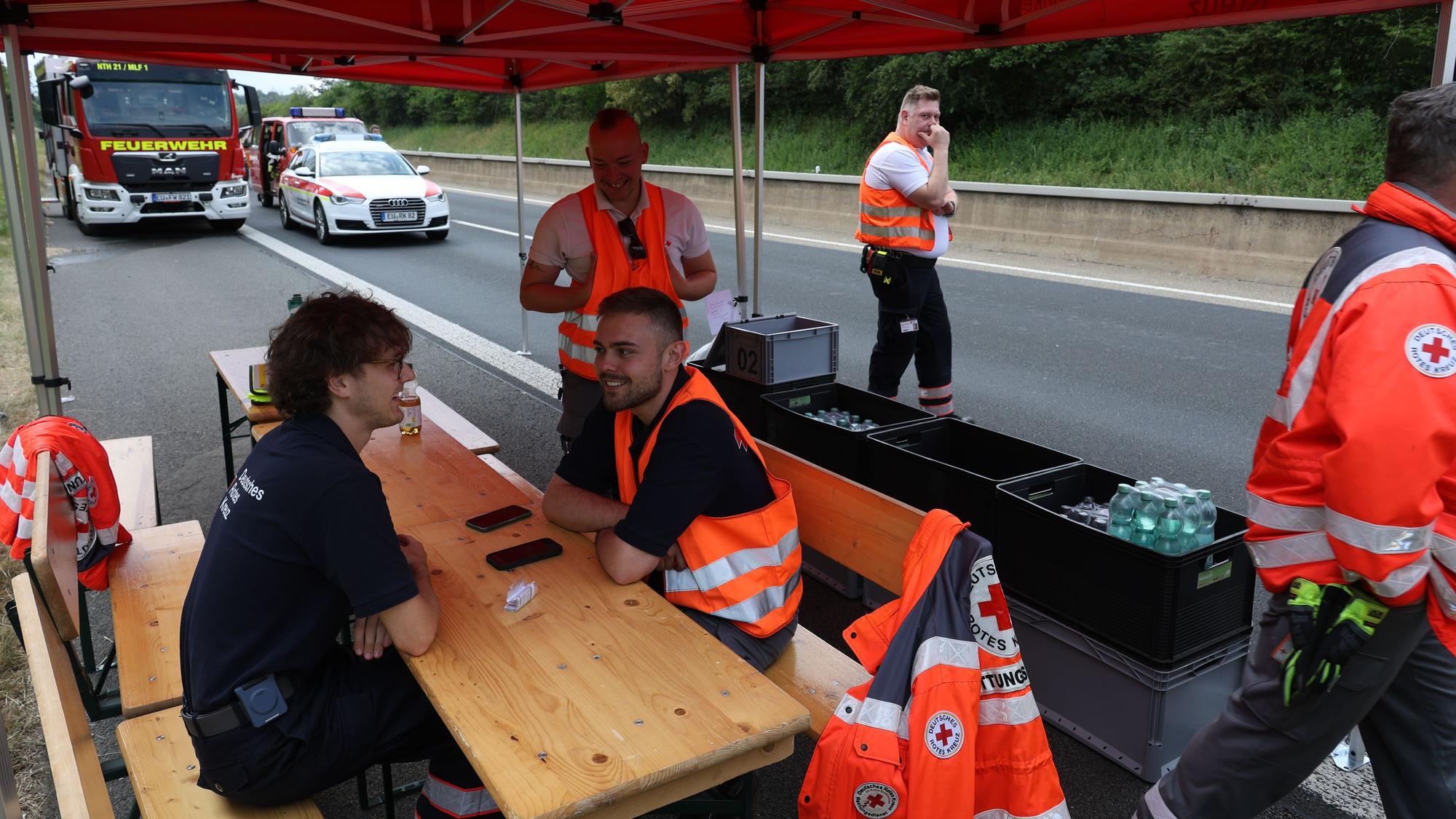 DRK-Helfer sitzen im Zelt auf der Autobahn vor Kisten mit Waserflaschen, im Hintergrund sind Einsatzfahrzeuge zu sehen.