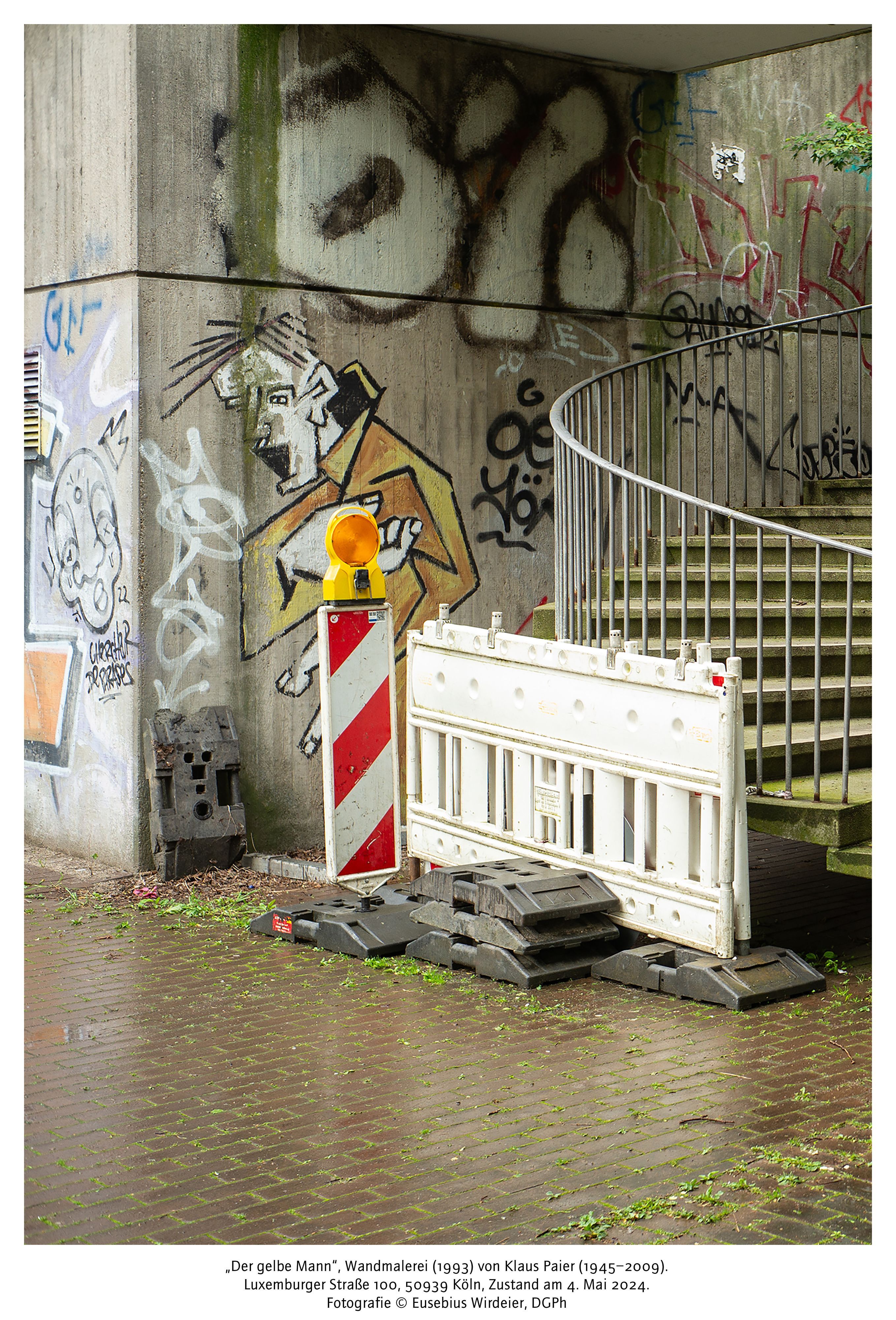 Hinter einem Bauzaun ist das Wandbild eines Gelben Mannes an einem Betonpfeiler zu sehen.