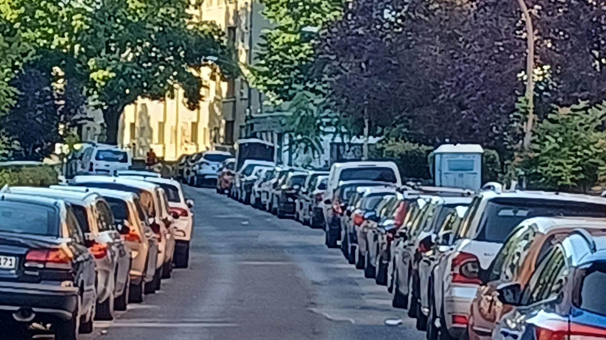 An einer mit Bäumen bestandenen Straße parken links und rechts Autos.