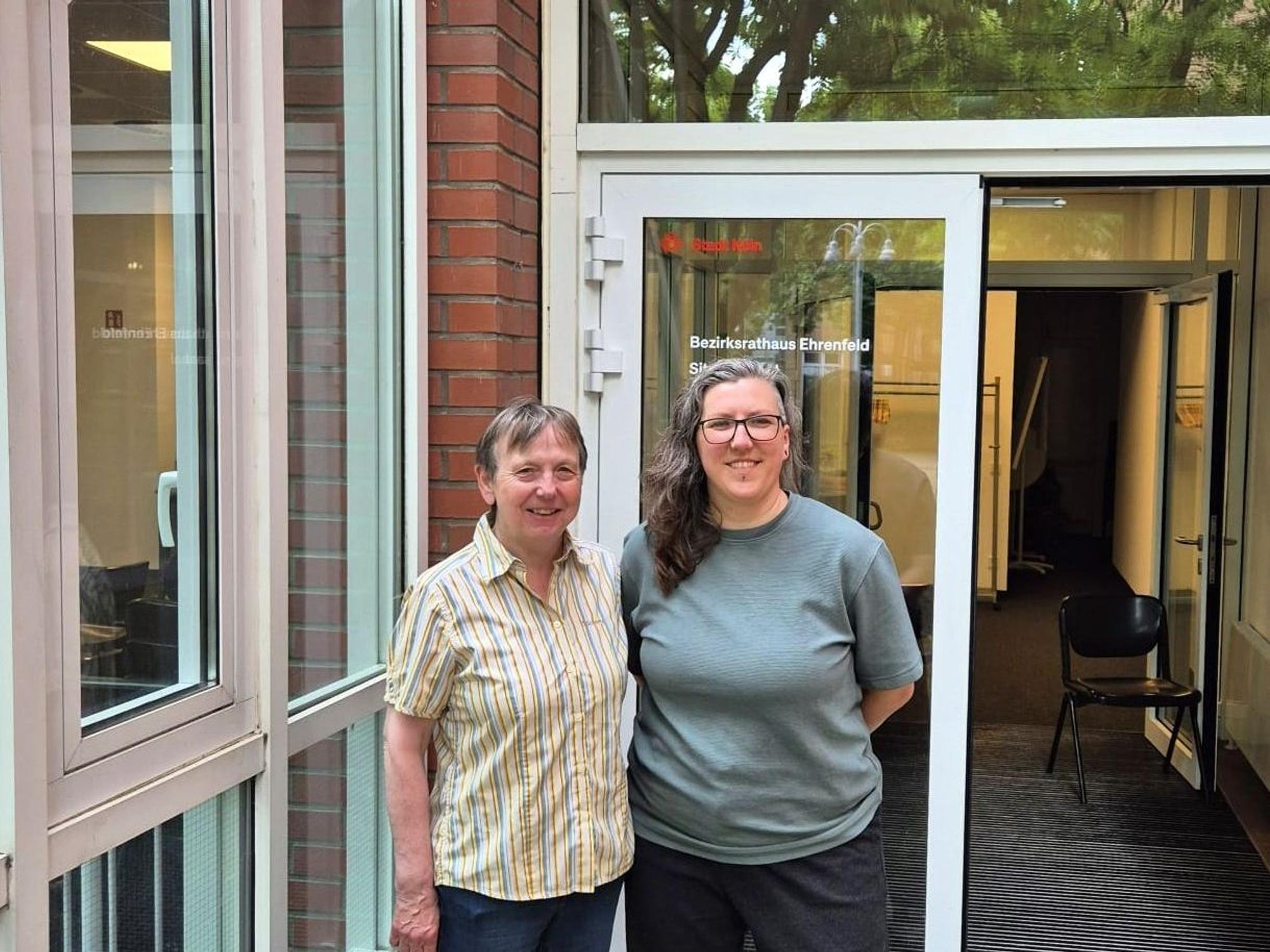 Eine Frau mit kurzem blondem Haar und eine mit langem grauen Haar stehen nebeneinander vor dem Eingang zum Bezirksrathaus in Ehrenfeld.