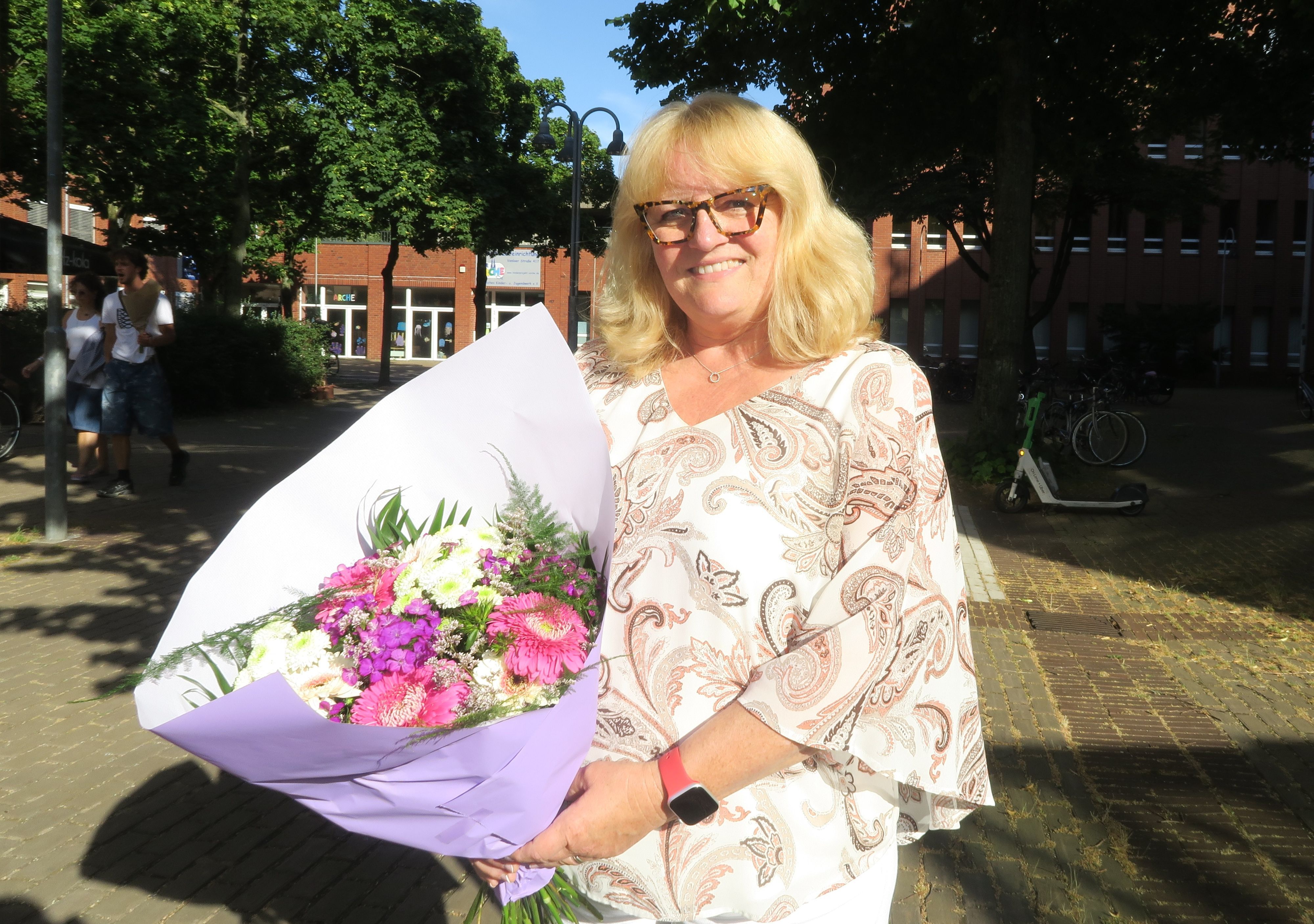 Eine Frau mit blondem Haar hält einen Blumenstrauß im Arm. Sie steht auf einem Platz in der Sonne.