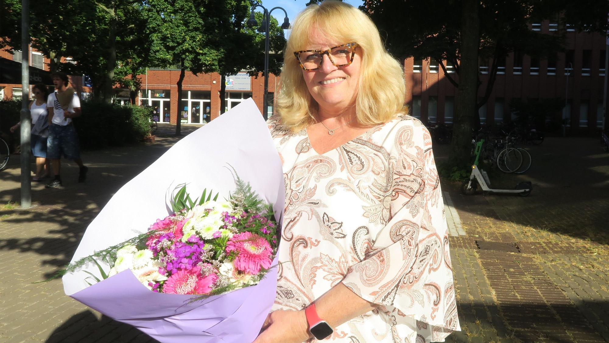 Eine Frau mit blondem Haar hält einen Blumenstrauß im Arm. Sie steht auf einem Platz in der Sonne.